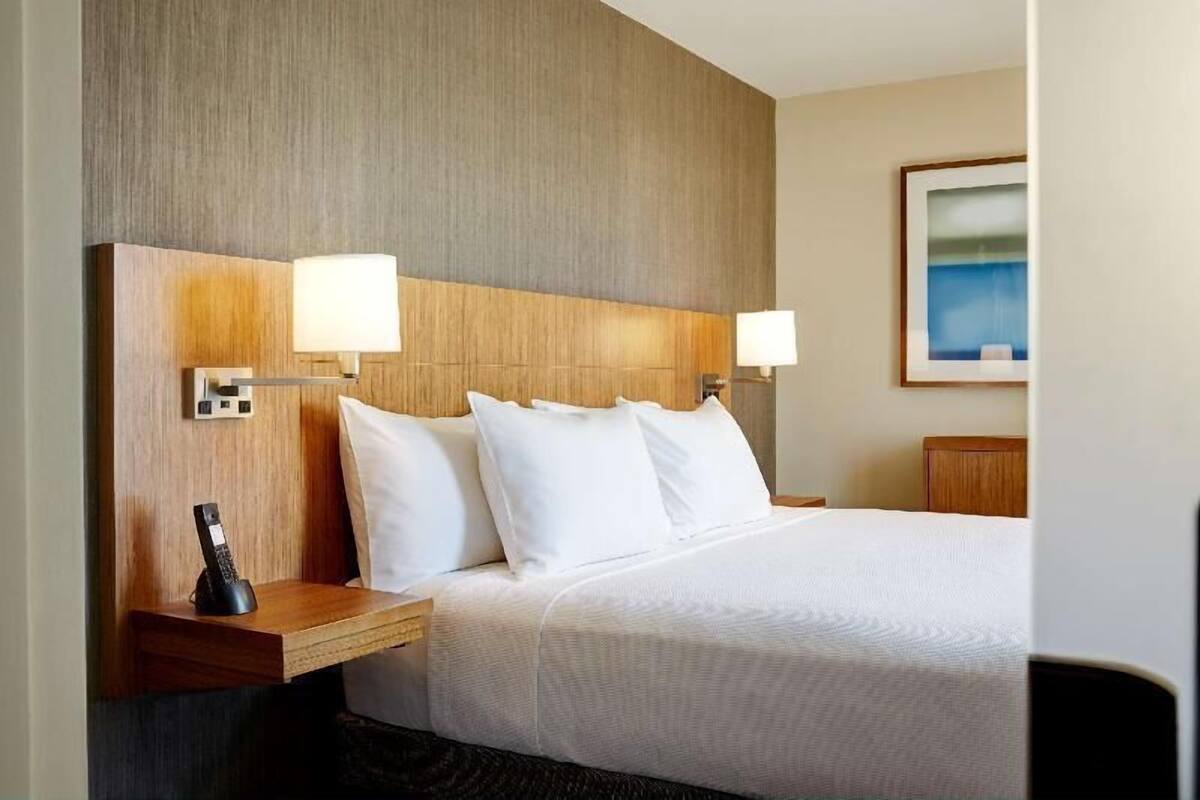 A hotel room with a bed, two bedside lamps, a phone on the nightstand, and a painting on the wall.