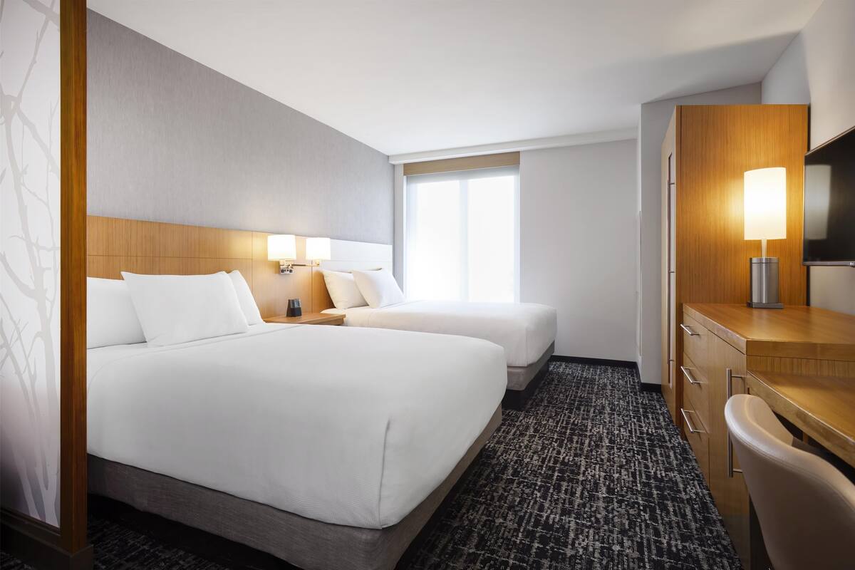 A hotel room with two beds, a desk, a chair, a TV, and a window.