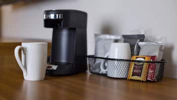 A coffee maker, a white mug, and a basket with coffee pods and a packet on a wooden table.