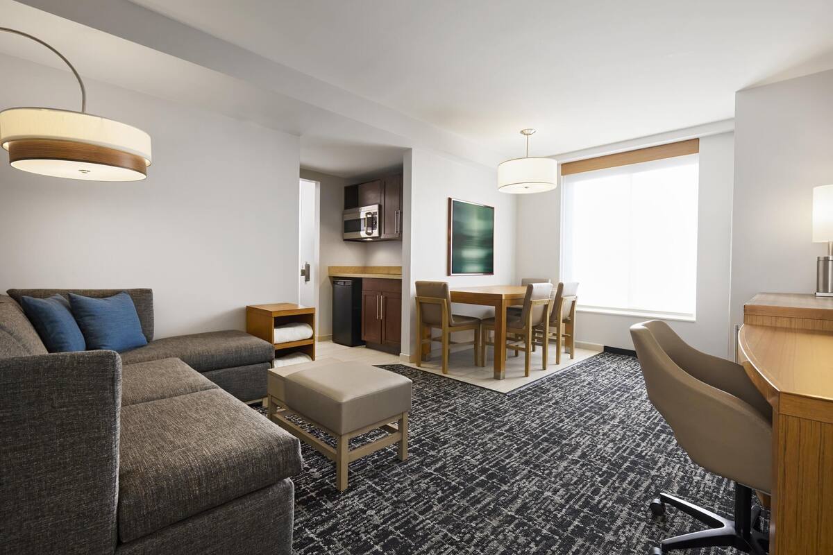 A modern hotel room with a sofa, ottoman, dining table, chairs, a desk, and a kitchen area.