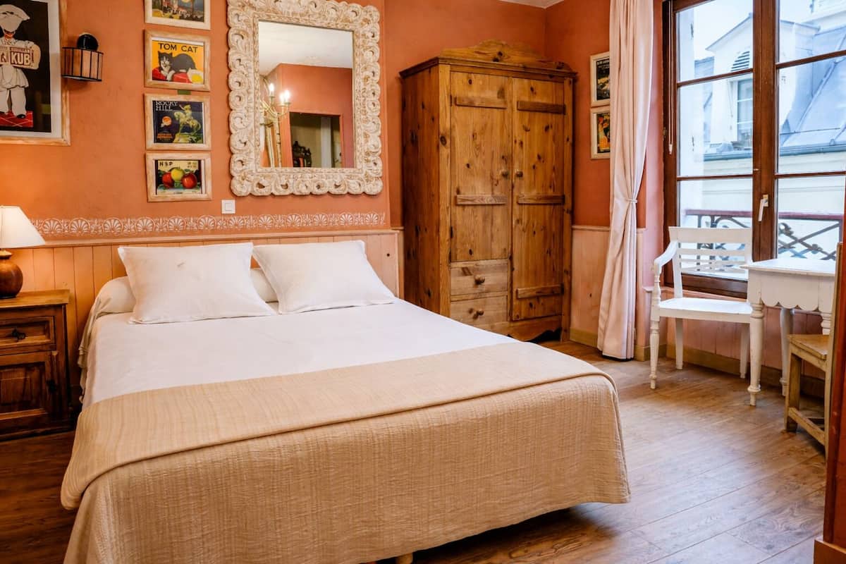 A bedroom with a large bed, a wooden wardrobe, a desk with a chair, and a window with a view of buildings.