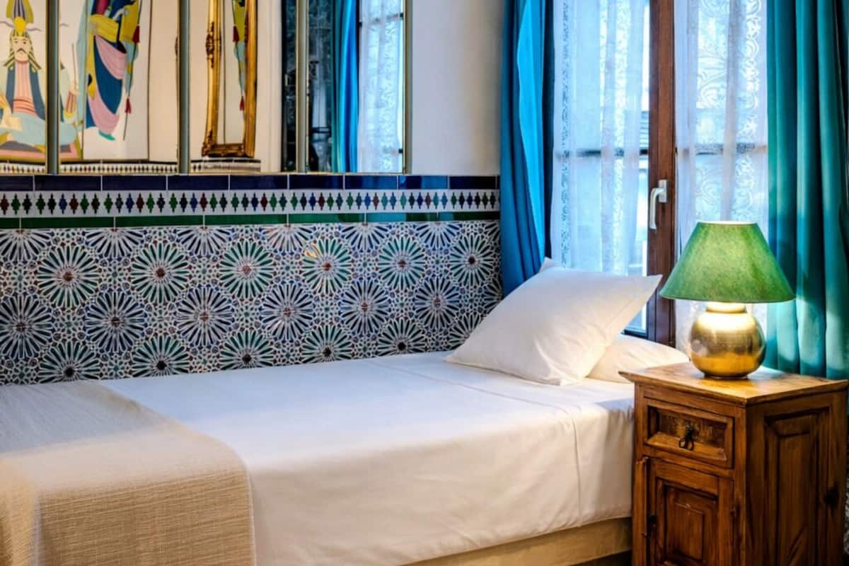 A room with a bed, a nightstand, a lamp, a window with curtains, and a wall with a decorative tile border.
