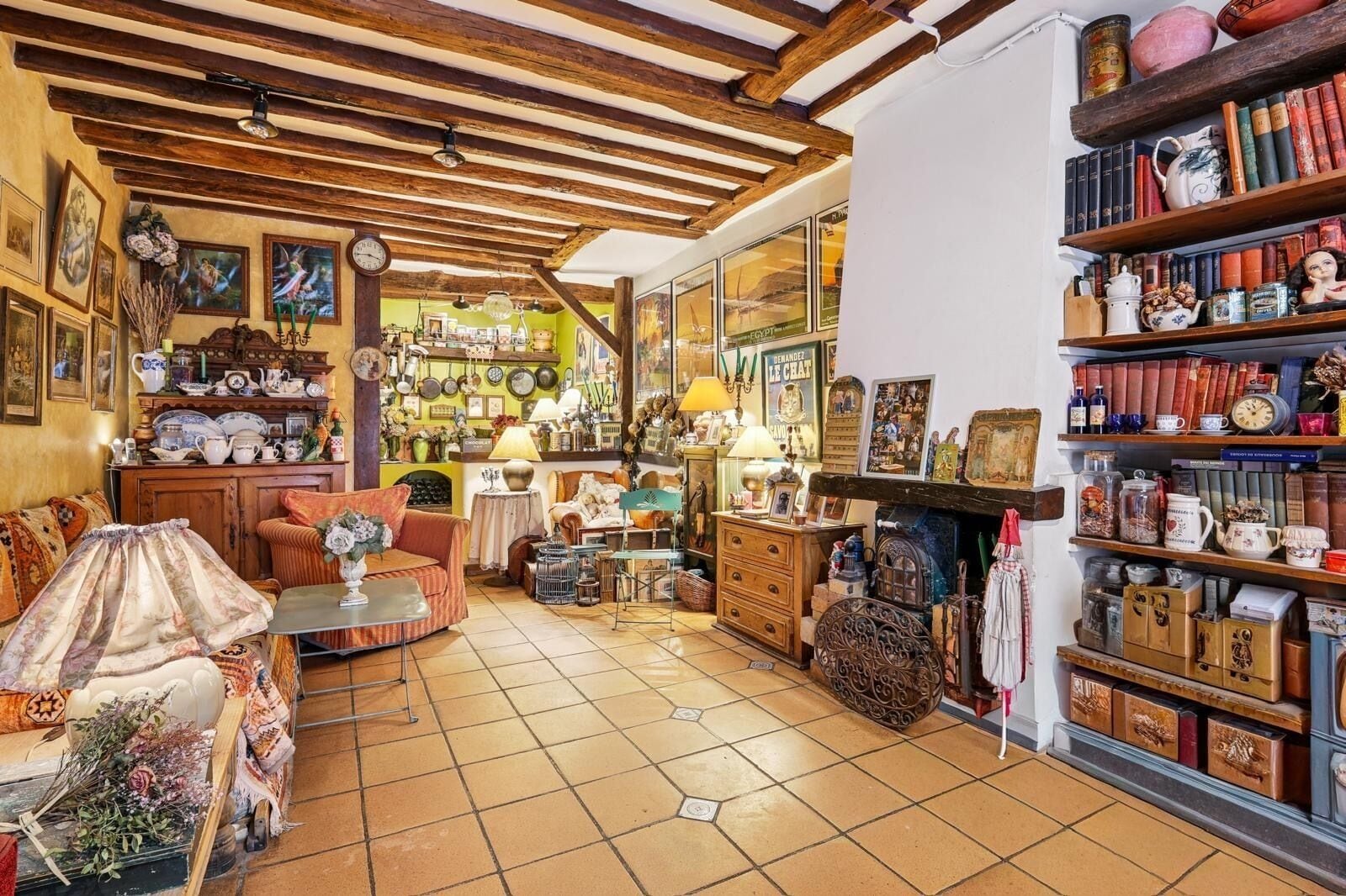Een knus interieur met een betegelde vloer, houten balken in het plafond, een bank, een tafel en planken vol boeken en decoratieve objecten.