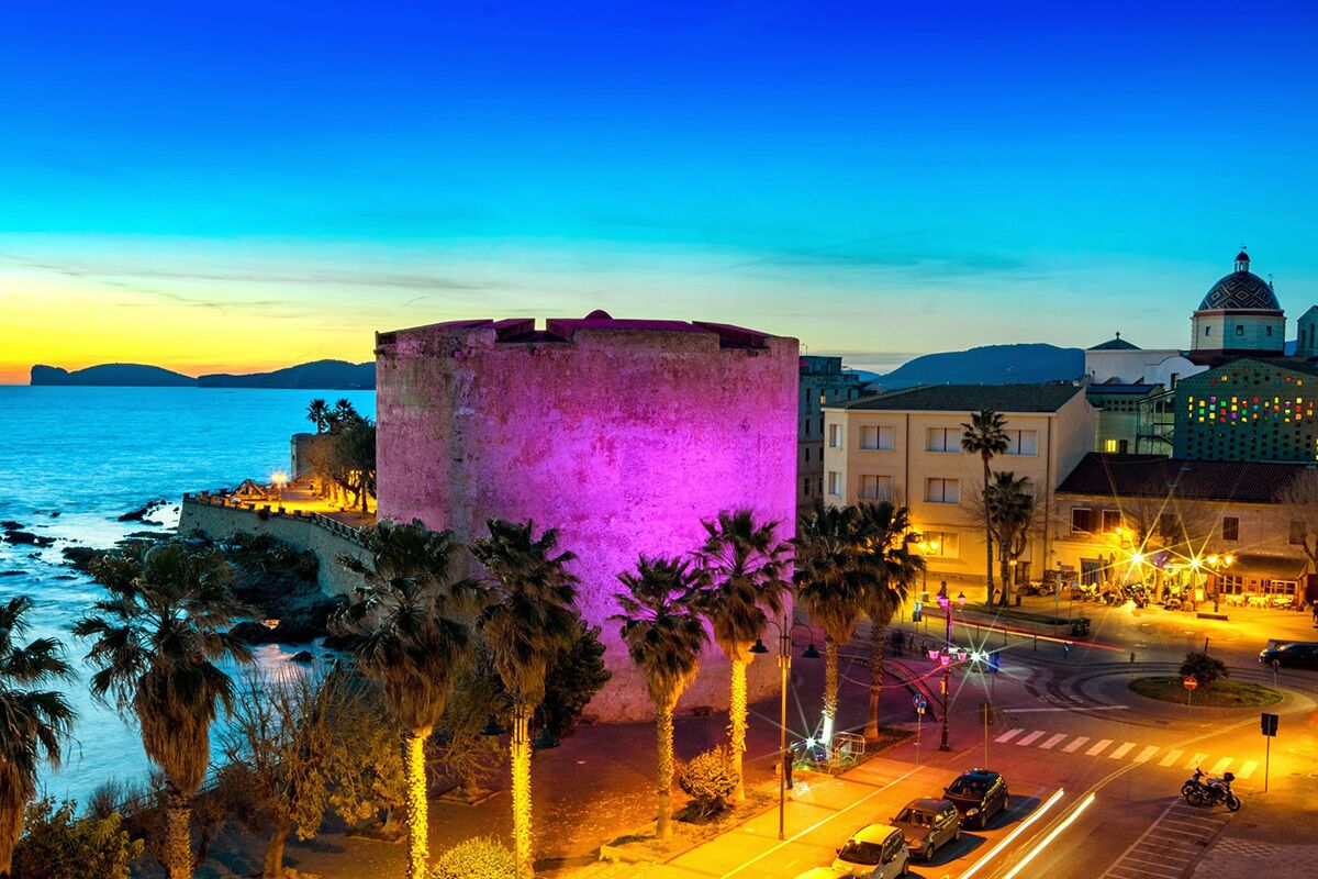 Un paisaje urbano costero con una destacada torre histórica, edificios modernos y una calle bien iluminada con palmeras.