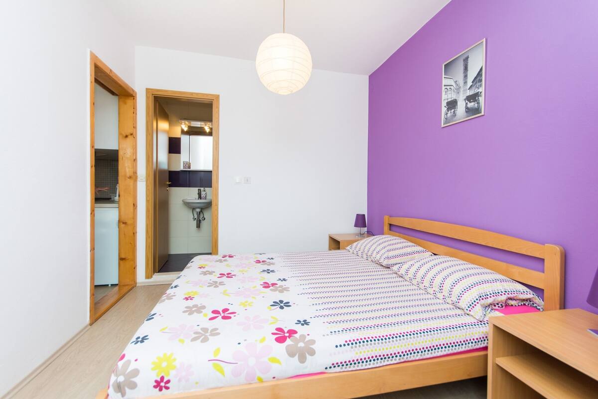 A bedroom with a bed, a wooden nightstand, a wall-mounted picture, and a door leading to a bathroom.