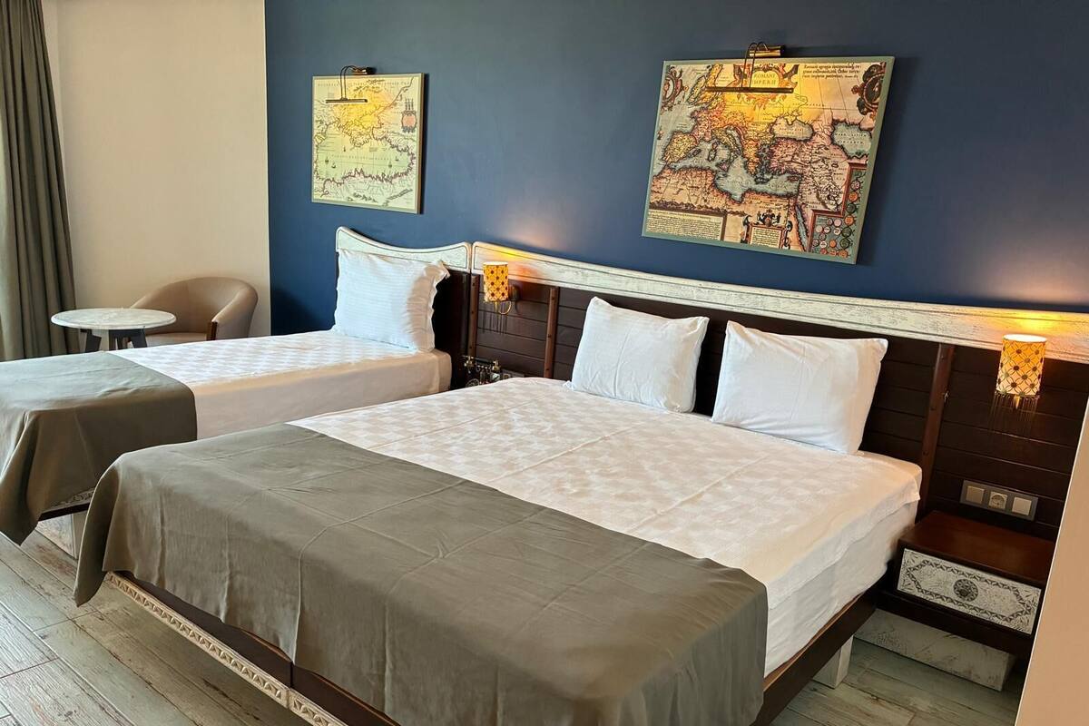 A hotel room with two beds, a chair, a small table, and two wall-mounted maps.