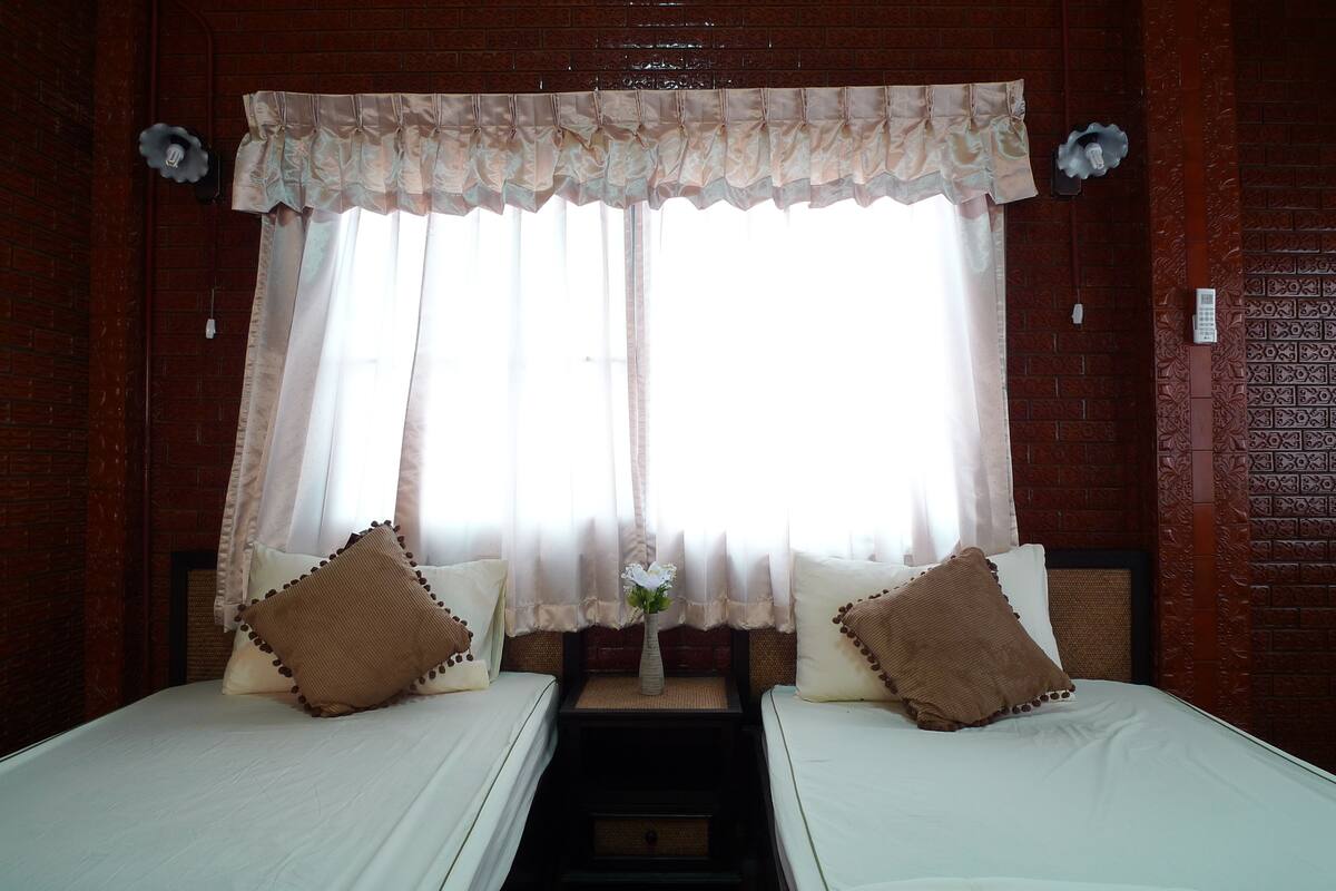 Una habitación con dos camas, una ventana con cortinas, un florero con flores y una pared de ladrillo.