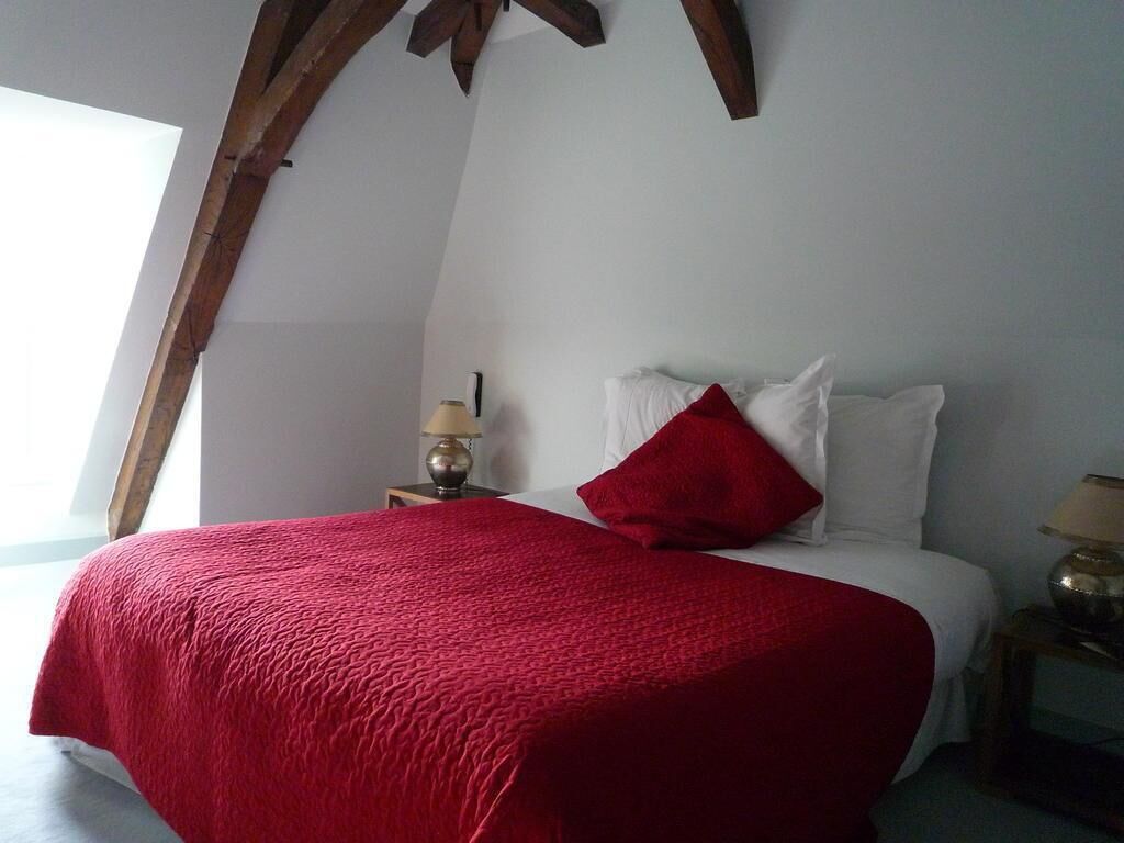 Een slaapkamer met een bed, een rode deken, witte kussens, een nachtkastje met een lamp en een houten balk.