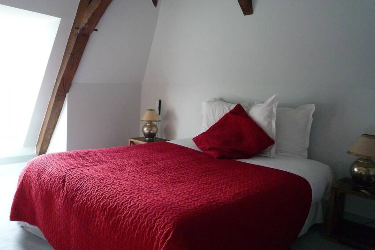 Een slaapkamer met een bed, een rode deken, witte kussens, een nachtkastje met een lamp en een houten balk.