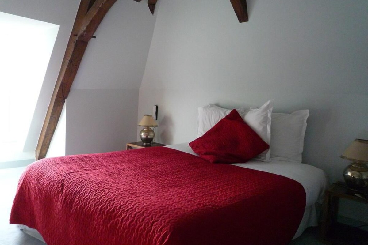 Een slaapkamer met een bed, een rode deken, witte kussens, een nachtkastje met een lamp en een houten balk.