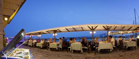 An outdoor dining area with tables and chairs, a fish display, and a view of the sea.