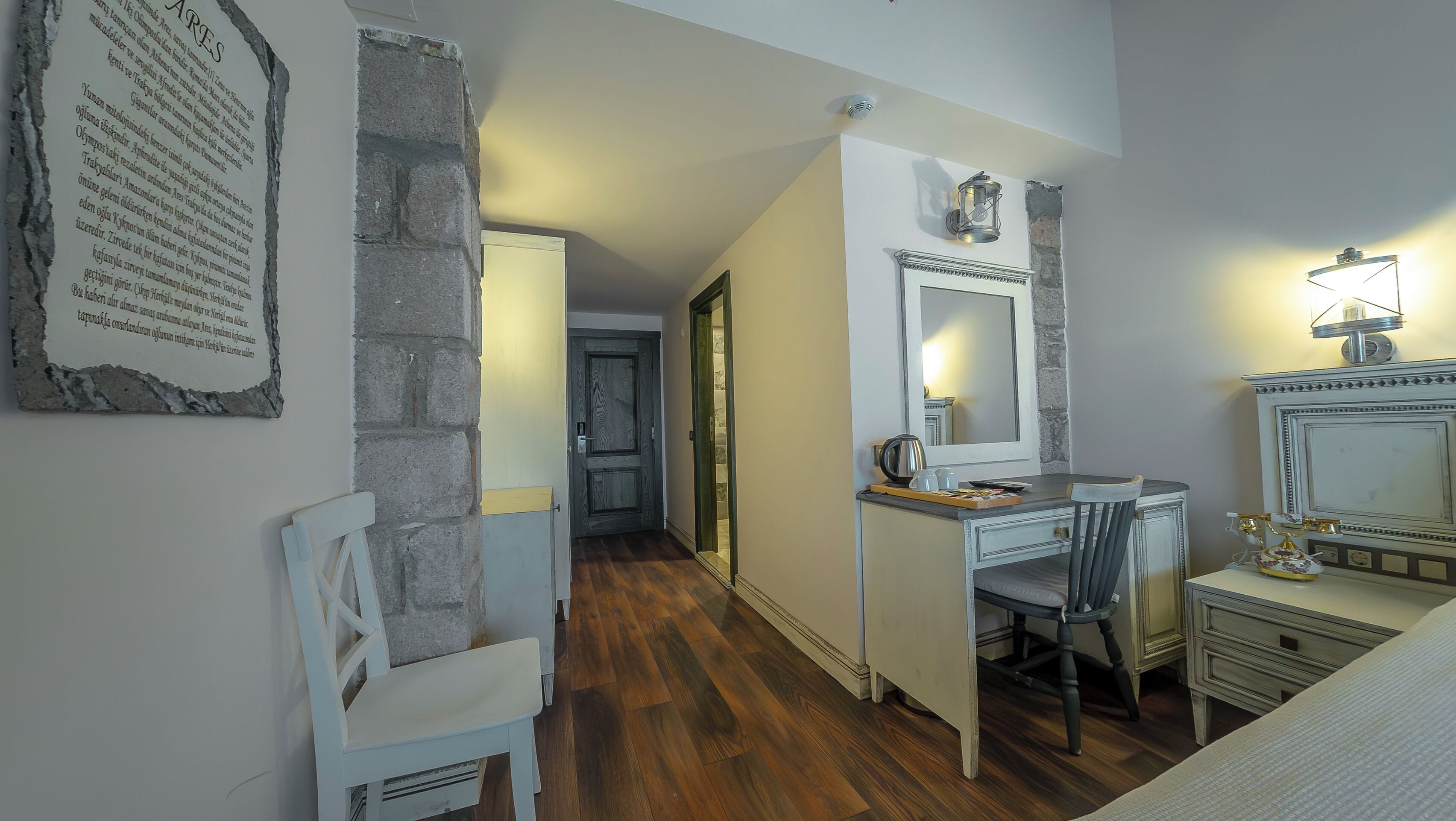 A hotel room with a wooden floor, a desk with a chair, a mirror, a dresser, and a white chair.