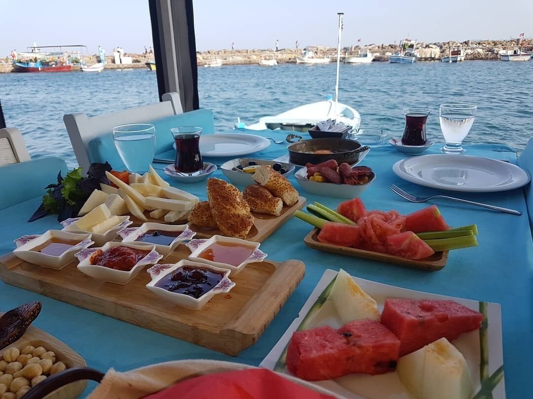 Una mesa servida con diversos alimentos y bebidas, con vista a barcos y una costa rocosa al fondo.