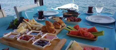 Una mesa servida con diversos alimentos y bebidas, con vista a barcos y una costa rocosa al fondo.