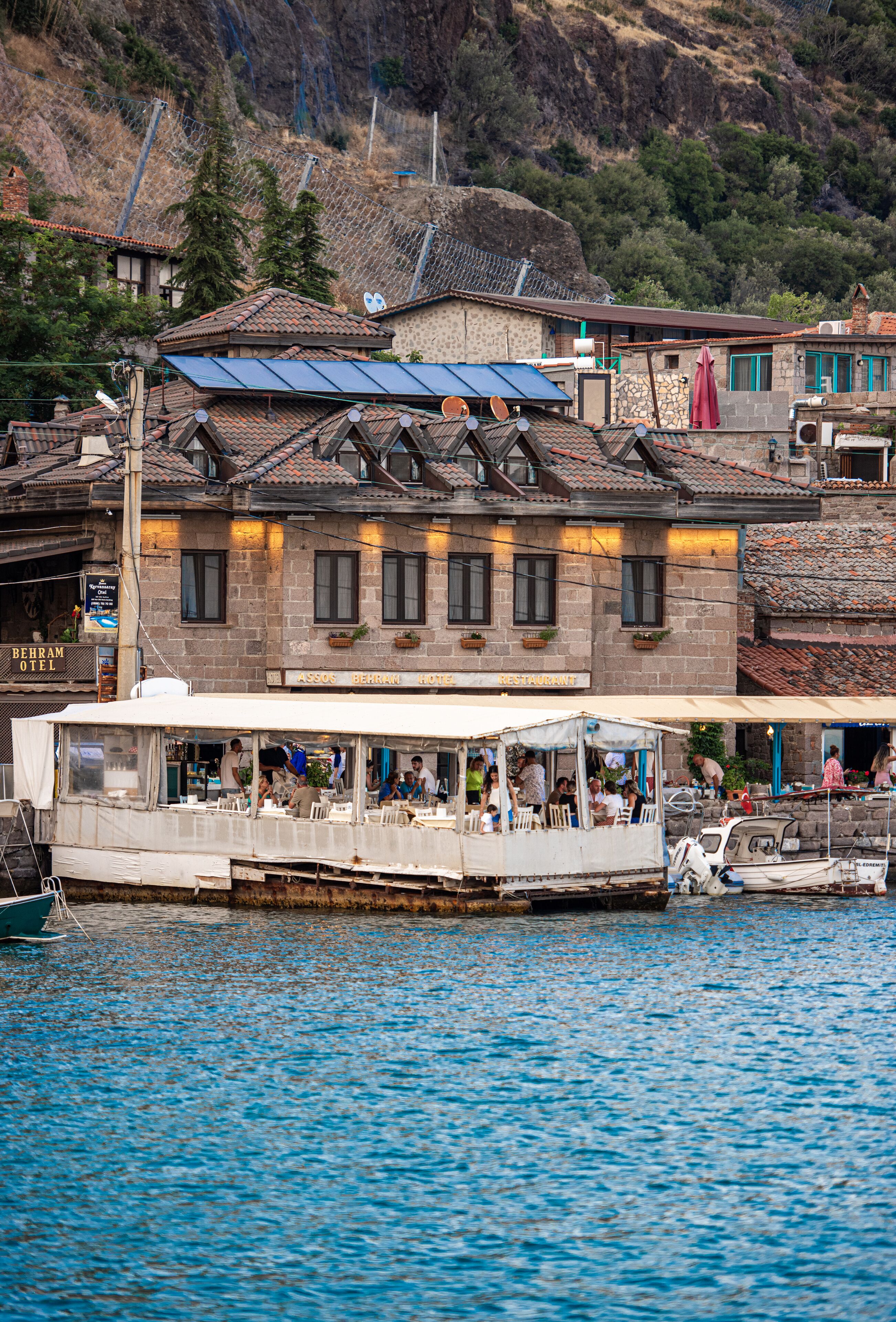 A boat with a covered dining area and a building with a sign that reads "BEHRAM HOTEL RESTAURANT" in the background.
