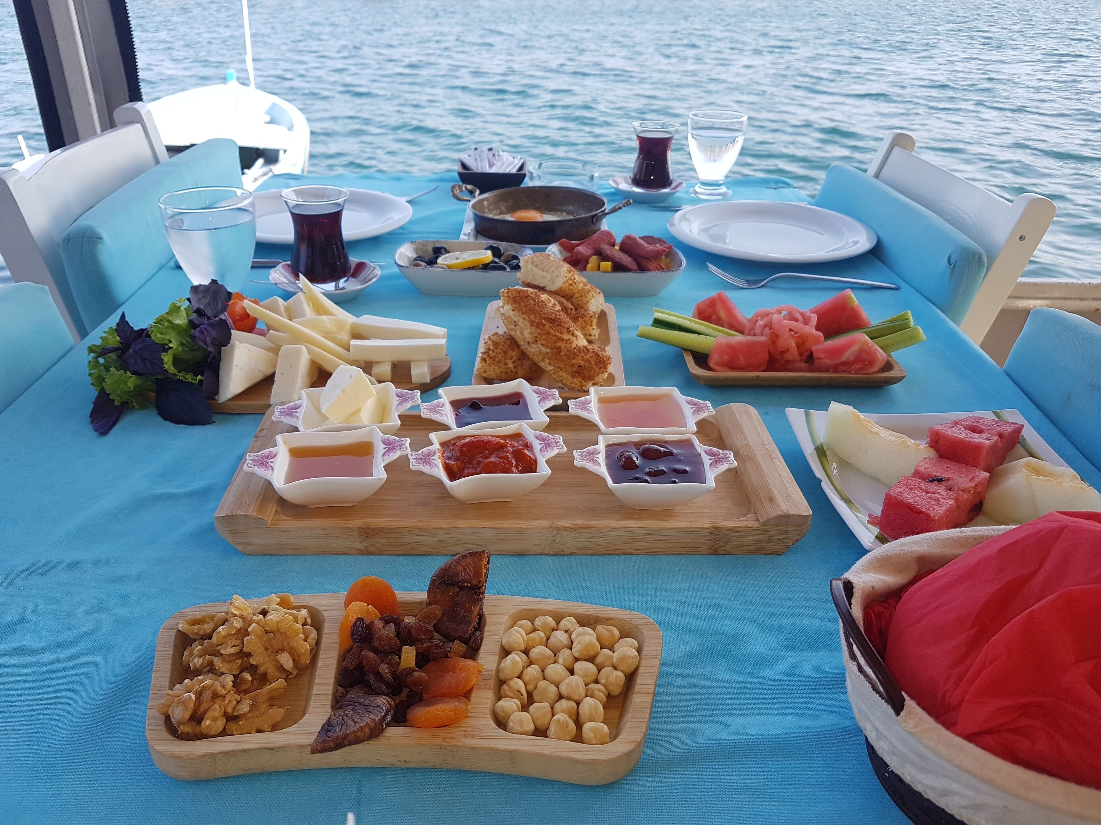 A table set with various foods and drinks, including cheese, fruits, and nuts, on a boat with a view of the water.