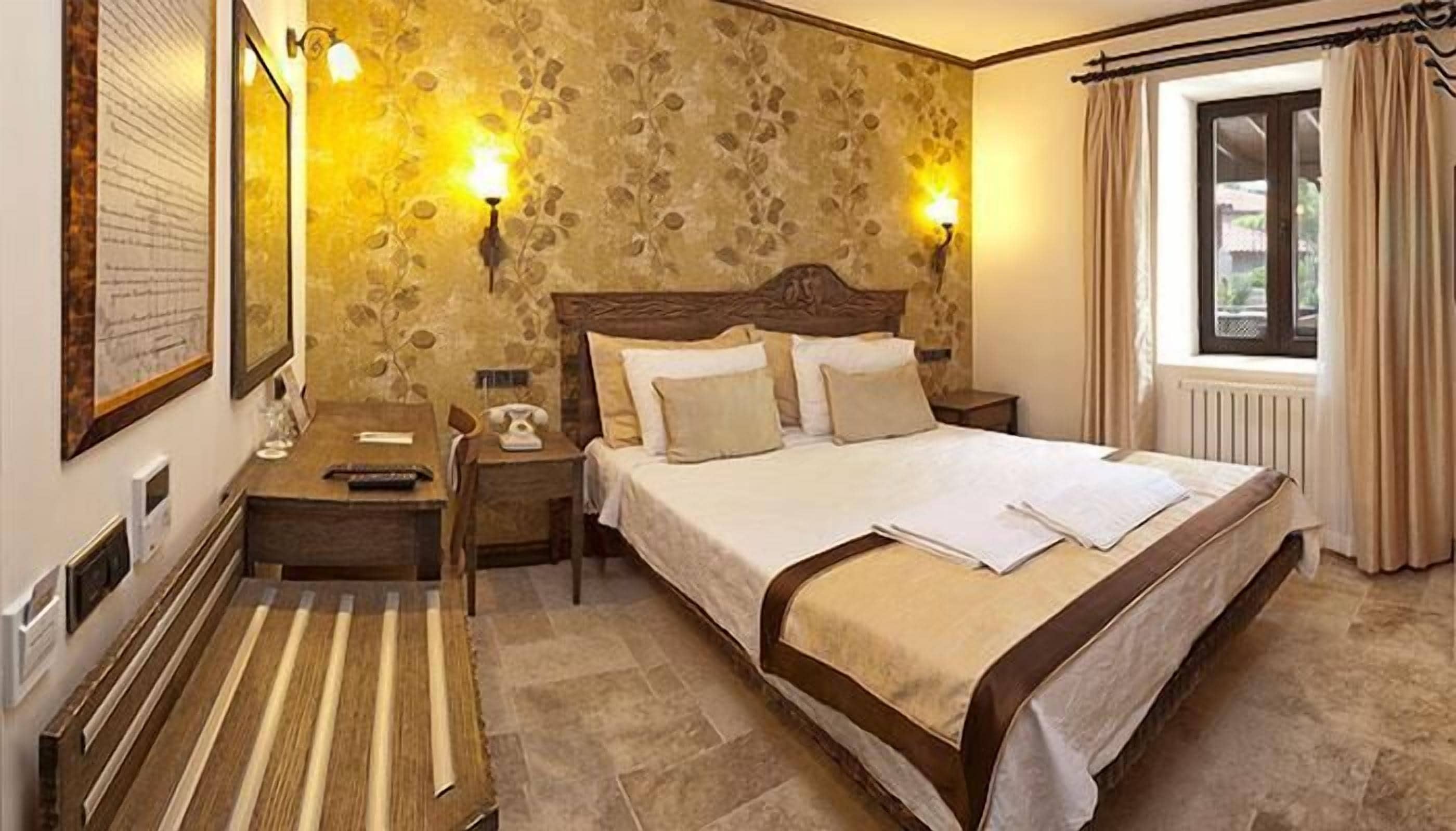 A hotel room with a stone-patterned wall, a wooden bed with white bedding, two bedside lamps, a nightstand with a telephone, and a window with curtains.