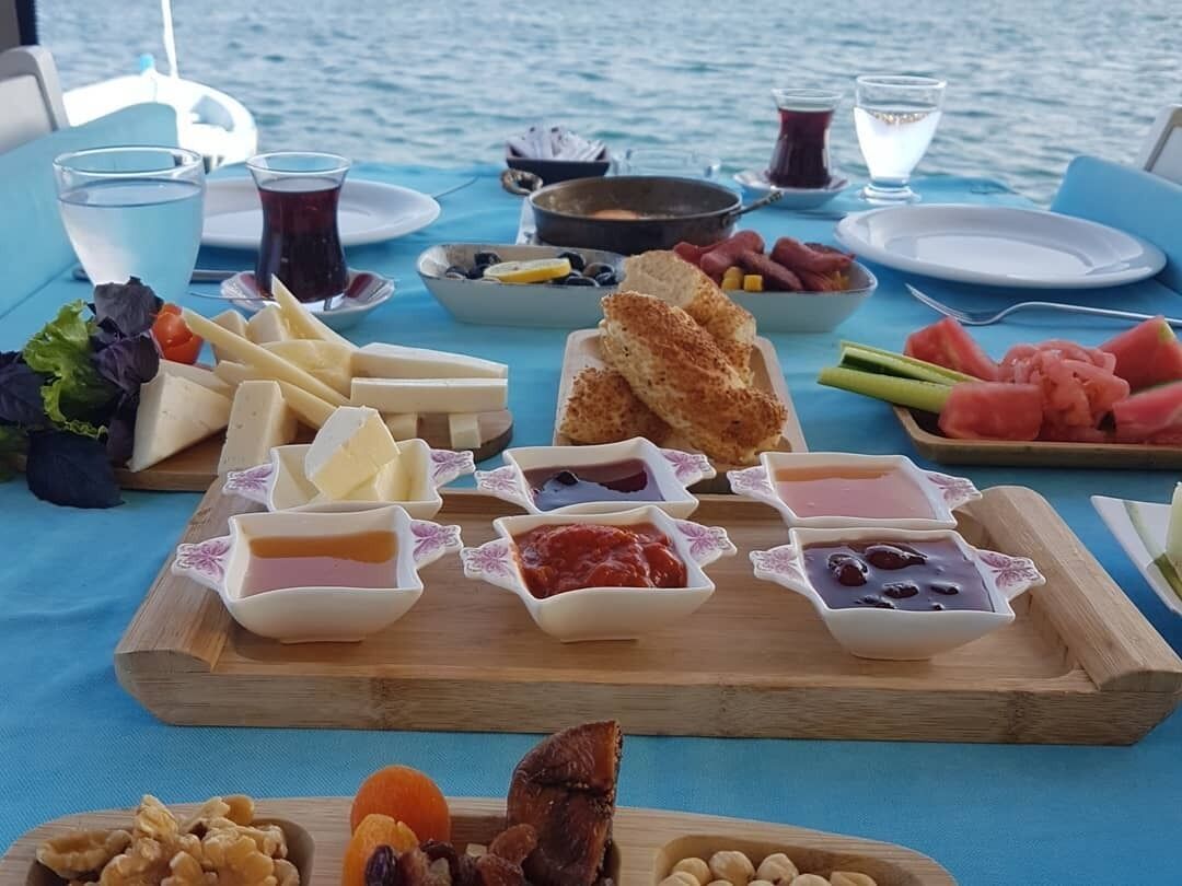 A table with various food items and drinks, including cheese, bread, fruits, and condiments, set against a backdrop of water.