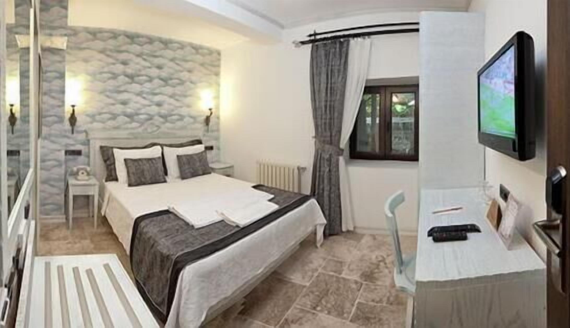 A bedroom with a stone wall, a bed with a white and gray bedspread, a nightstand, a TV on a stand, and a window with curtains.