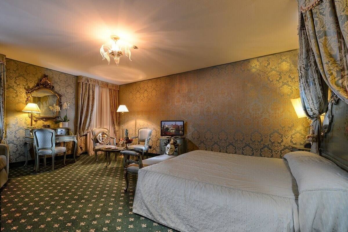 A hotel room with a large bed, a desk with a chair, a television, and a chandelier.