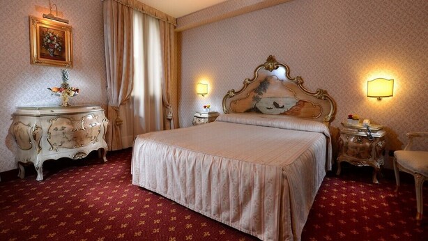 Une chambre d’hôtel avec un grand lit, une tête de lit décorée, deux tables de chevet avec des lampes, une commode et un miroir.