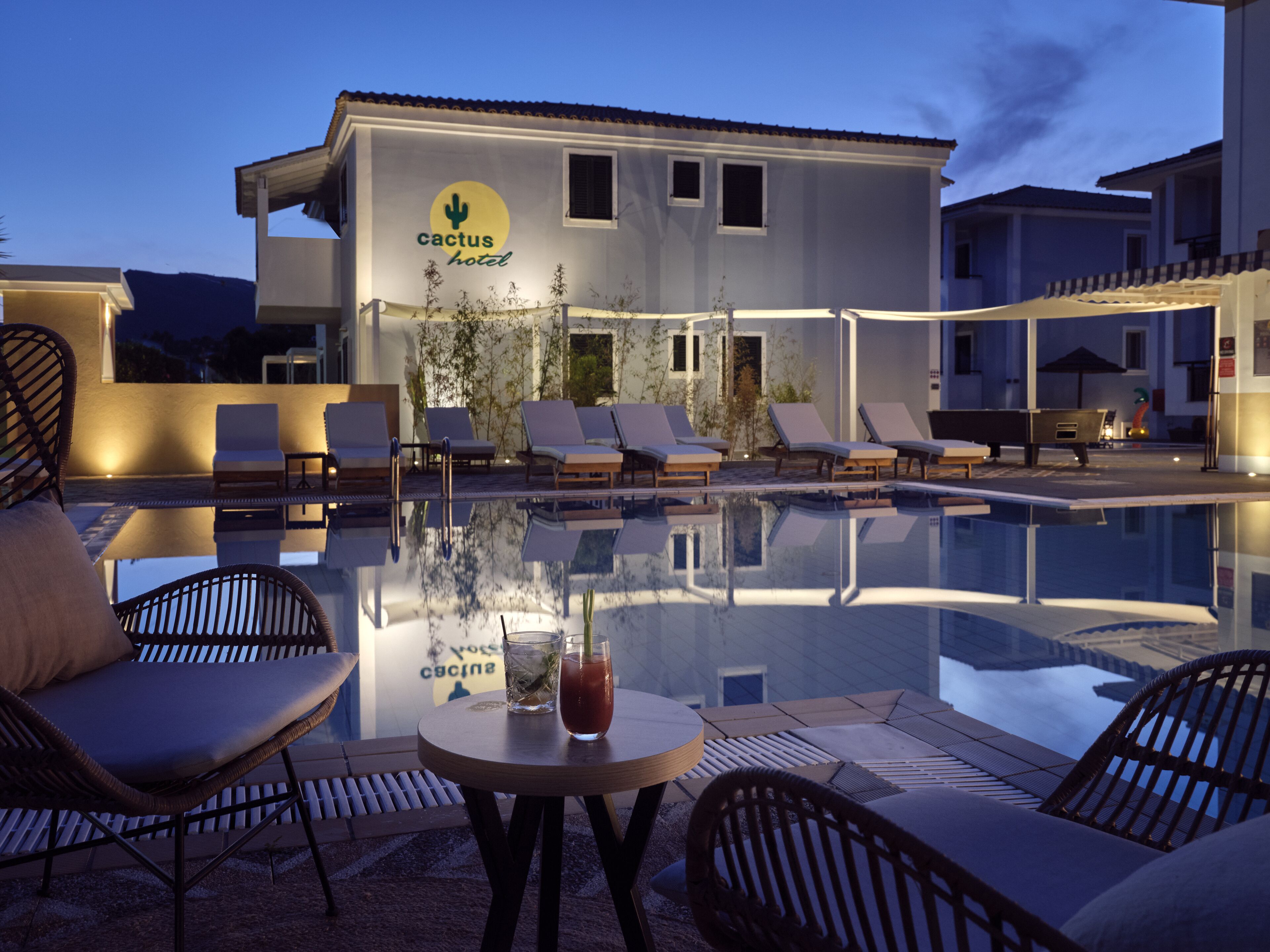 Zona de piscina con sillones de descanso y una mesa, un edificio con el logo de un cactus y una piscina cristalina que refleja el entorno.