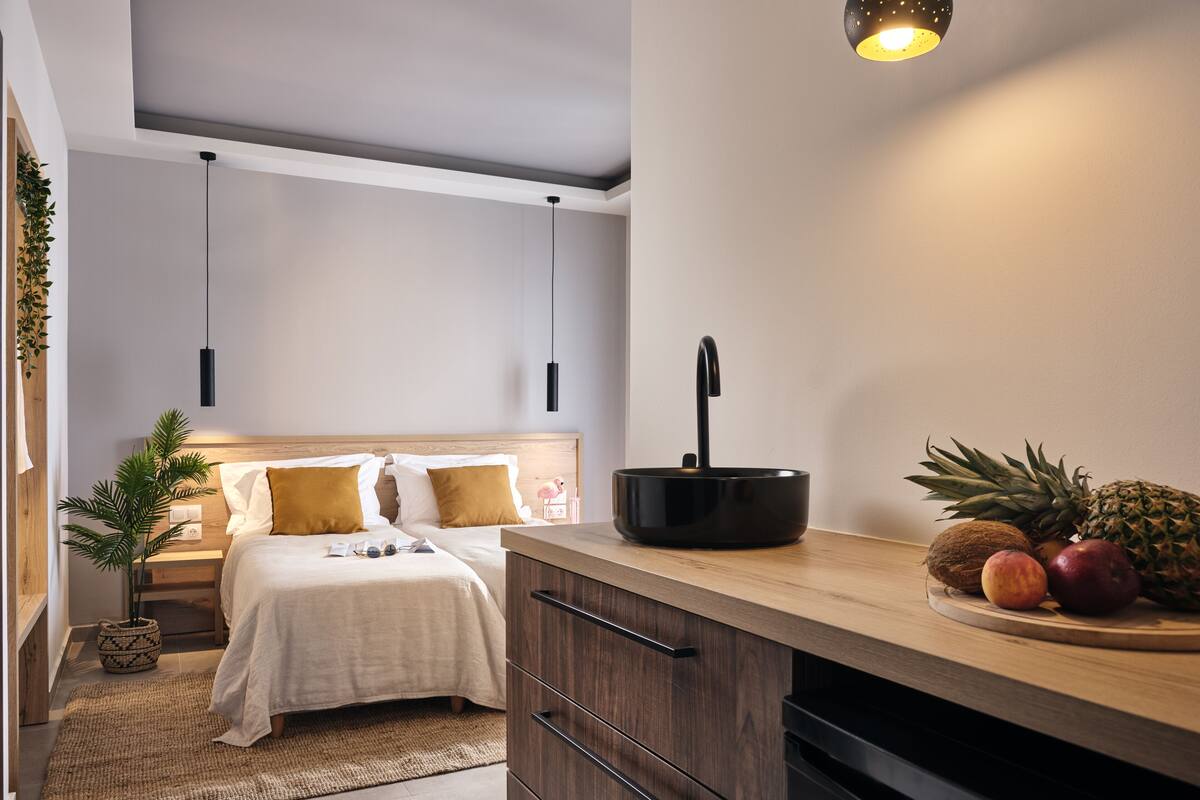 Una habitación de hotel moderna con cama, mesita de noche y una pequeña cocina con frutero.