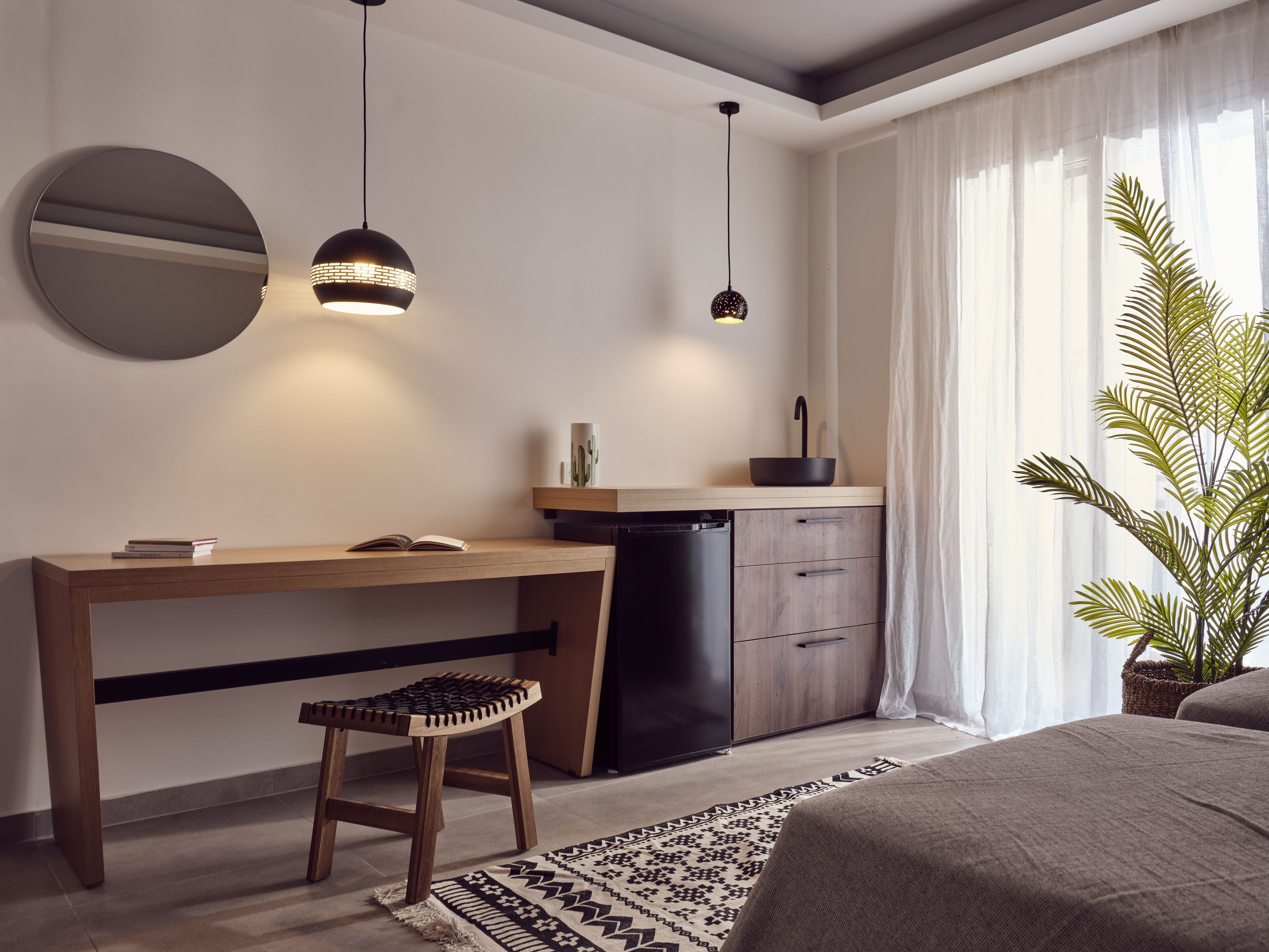 Een moderne slaapkamer met een houten bureau, een zwarte wastafel, een ronde spiegel en een kamerplant.