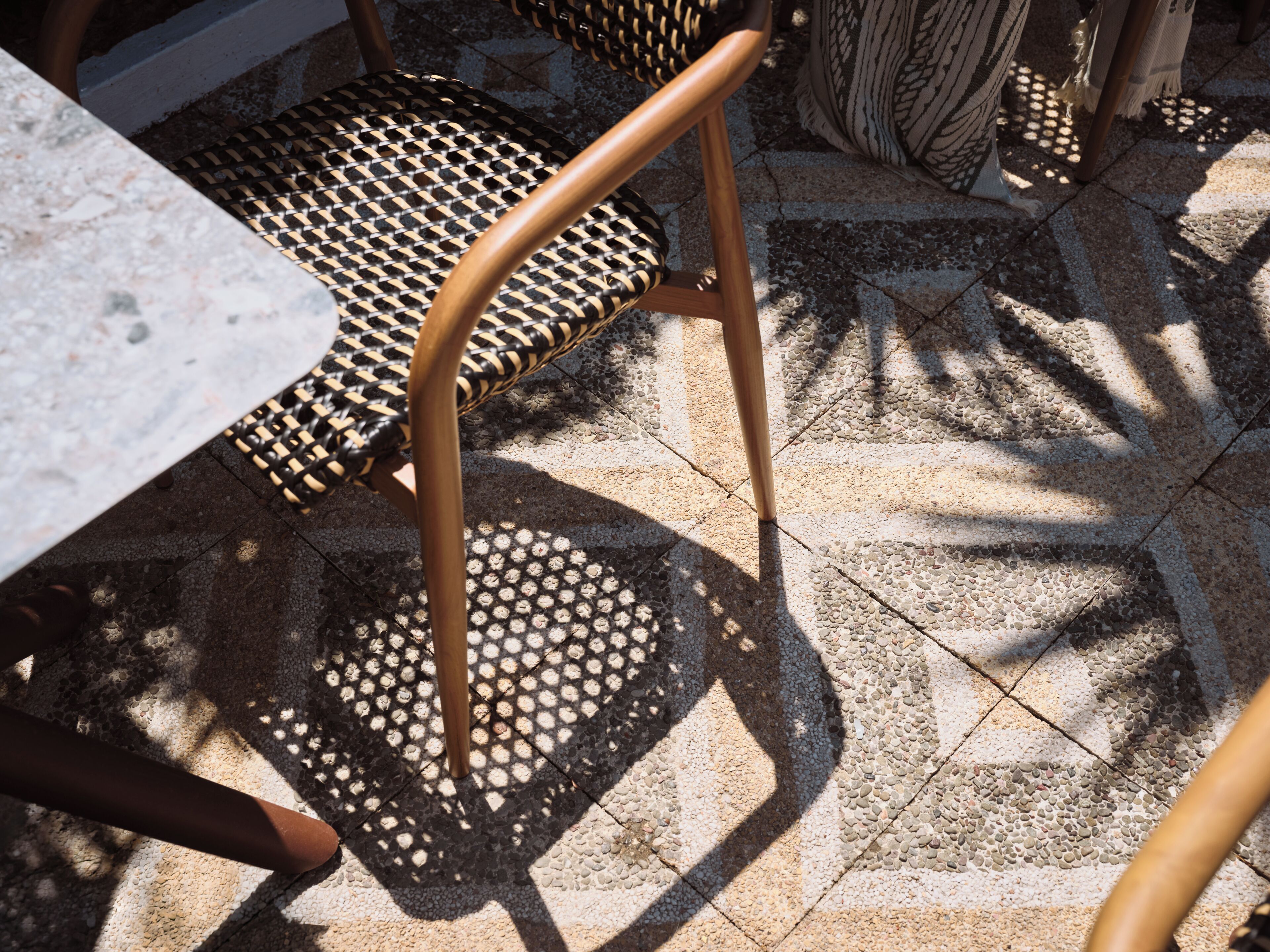 A chair with a woven seat and metal legs, a table with a stone top, and a patterned floor with shadows cast by nearby objects.