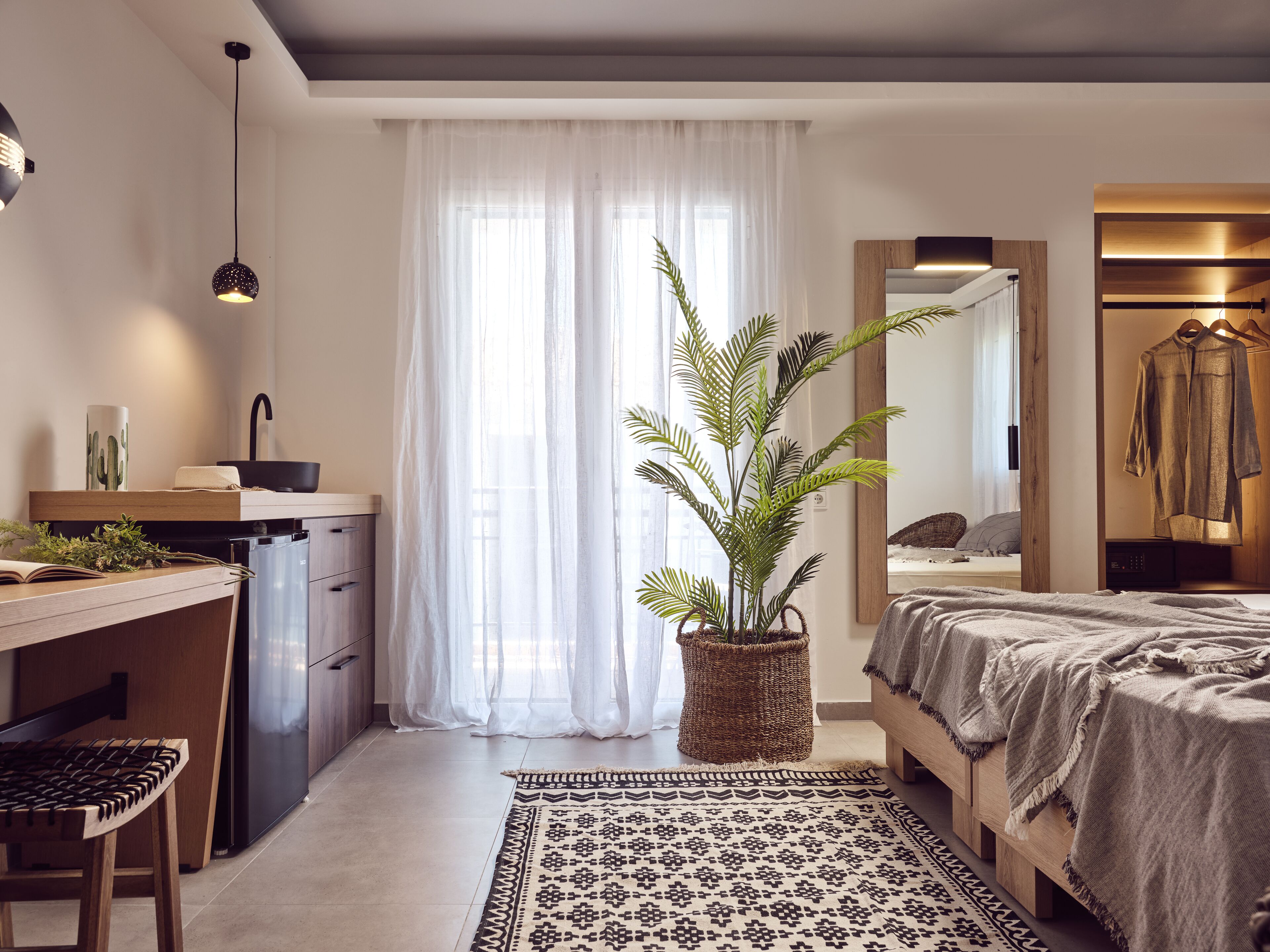 A modern bedroom with a bed, bedside table, and a large mirror. There is a potted plant in the corner and a window with sheer curtains.