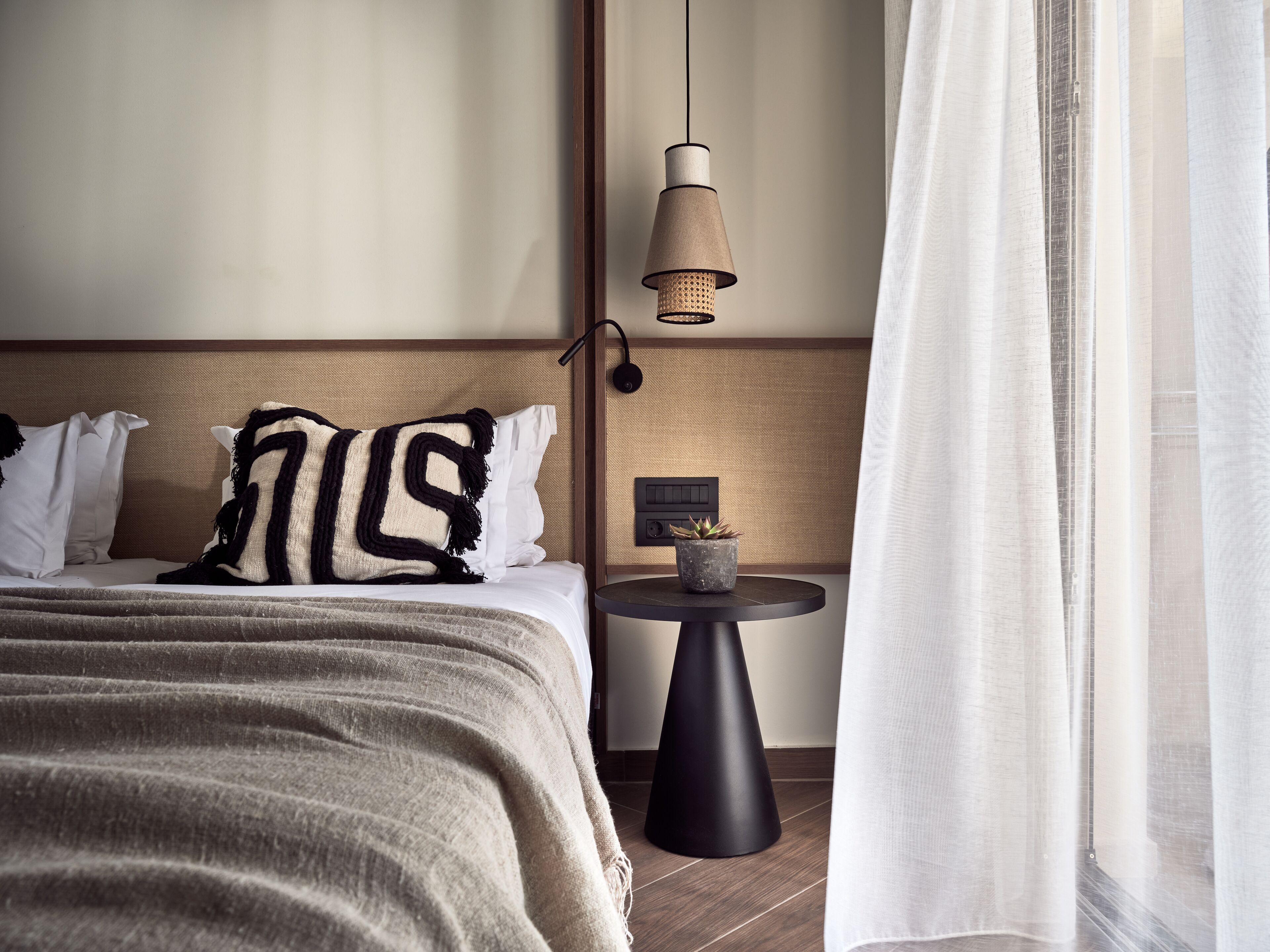 Un lit bien fait, recouvert d’une couverture à rayures et agrémenté de deux oreillers, une petite table d’appoint ronde et noire sur laquelle est posée une plante en pot, un luminaire suspendu et une fenêtre ornée de rideaux légers.