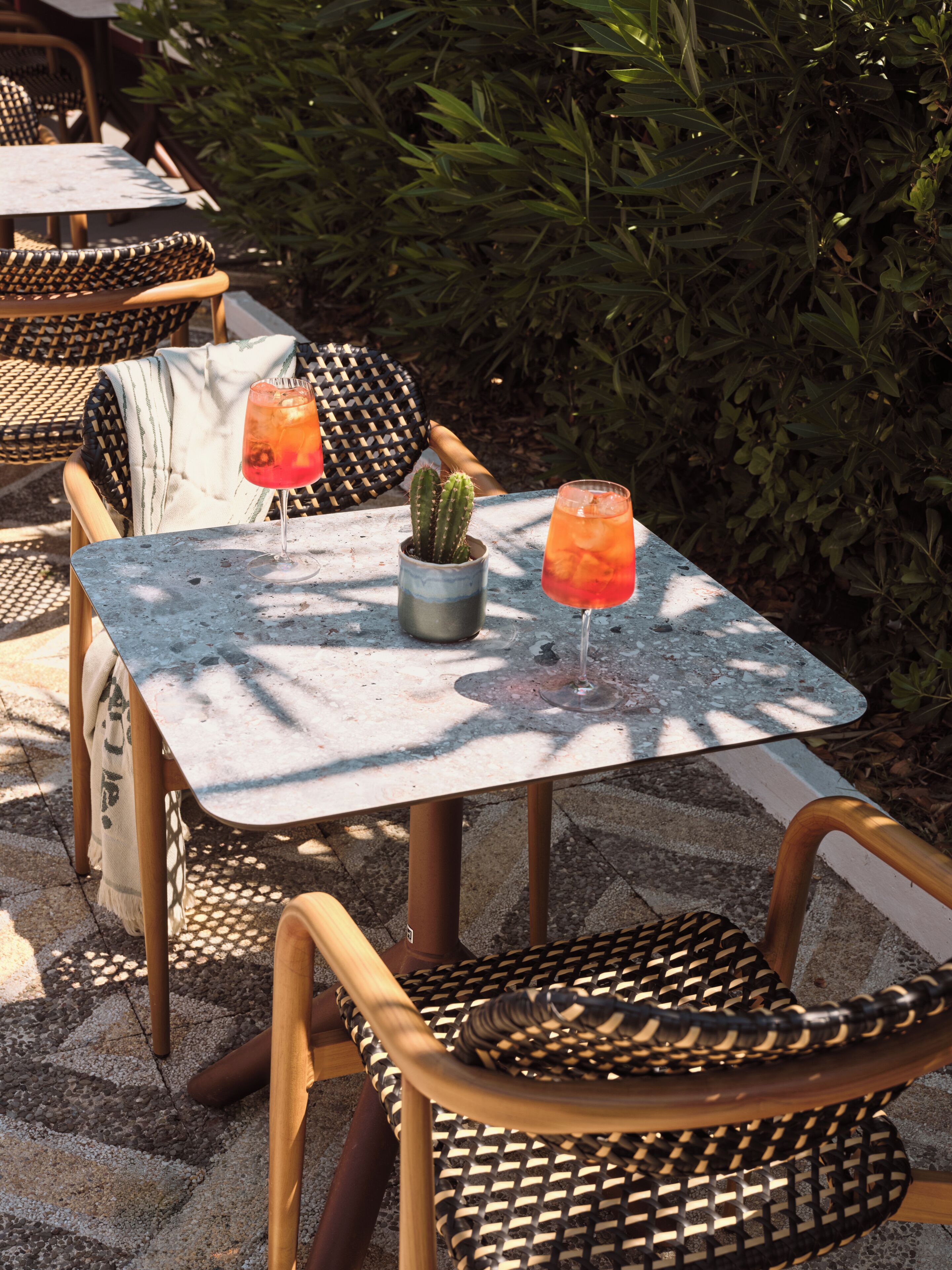 Mobiliario exterior con sillas de mimbre y una mesa, dos bebidas y una maceta con cactus.