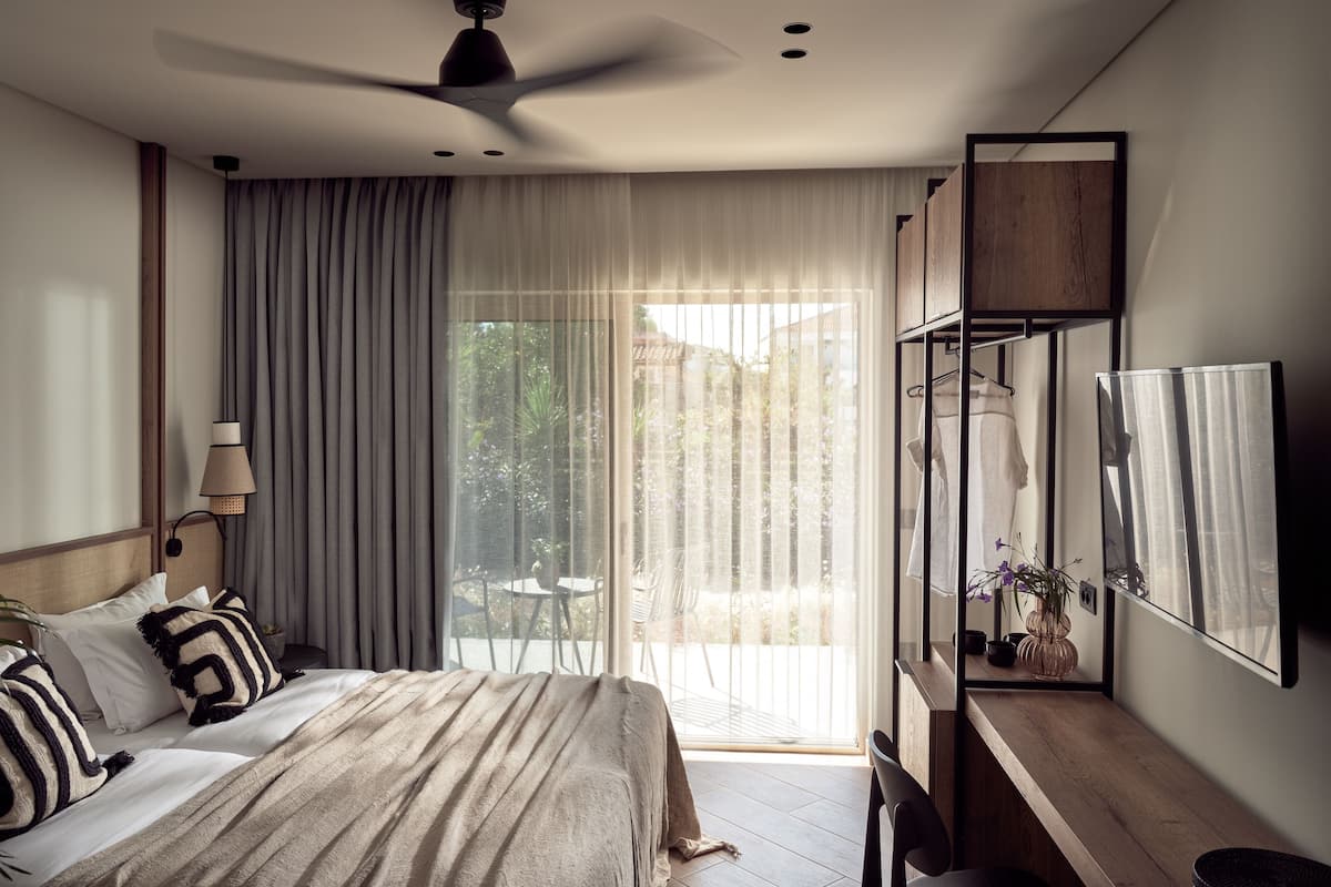 Un dormitorio moderno con cama, ventilador de techo, ventana grande con cortinas traslúcidas, mesita de noche de madera y espejo.