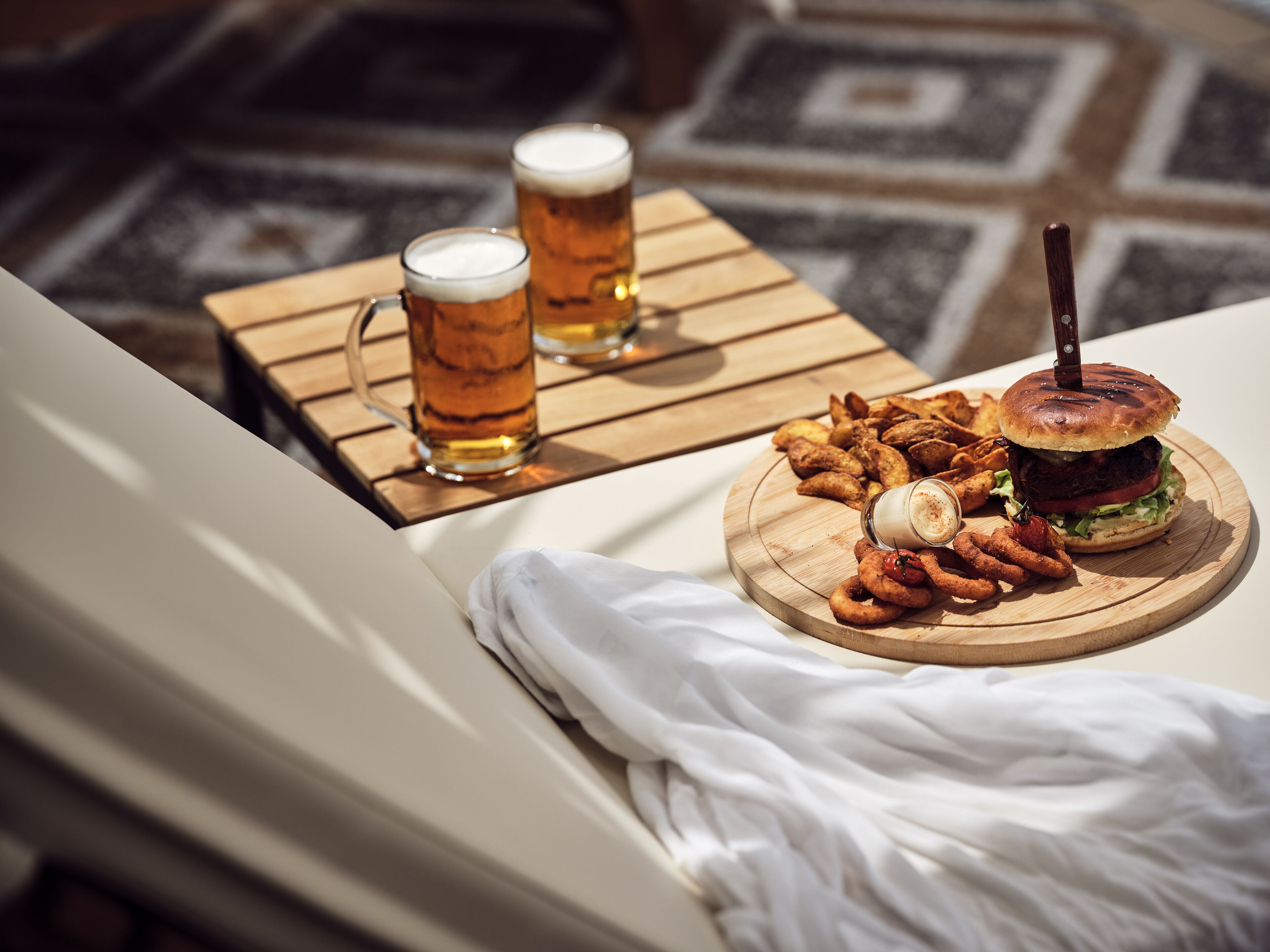 A dining table with a burger, onion rings, and two beers on a wooden board.