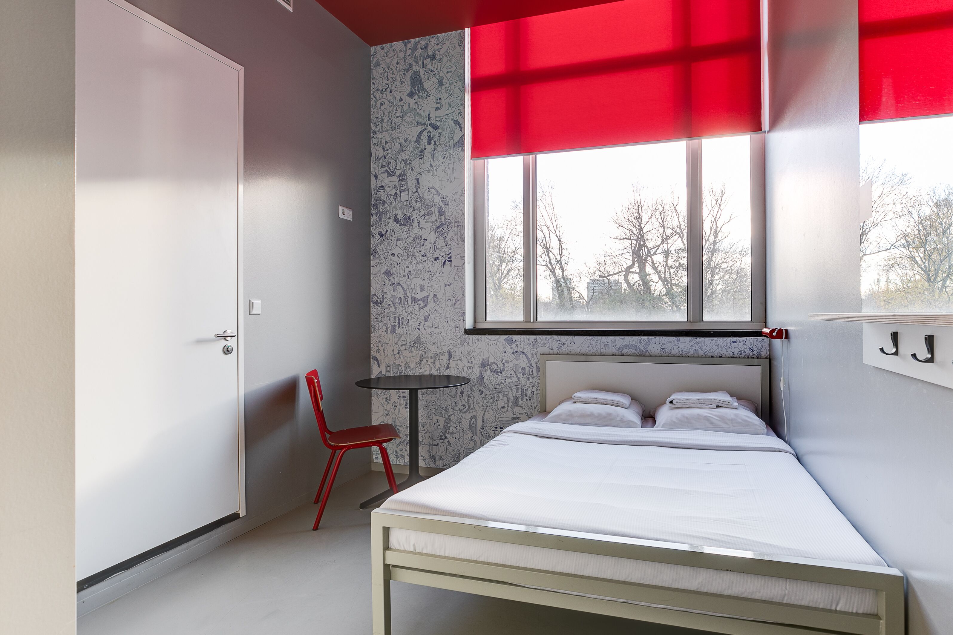 Een slaapkamer met een bed, een rode stoel, een tafeltje en een raam met een rood klepje.
