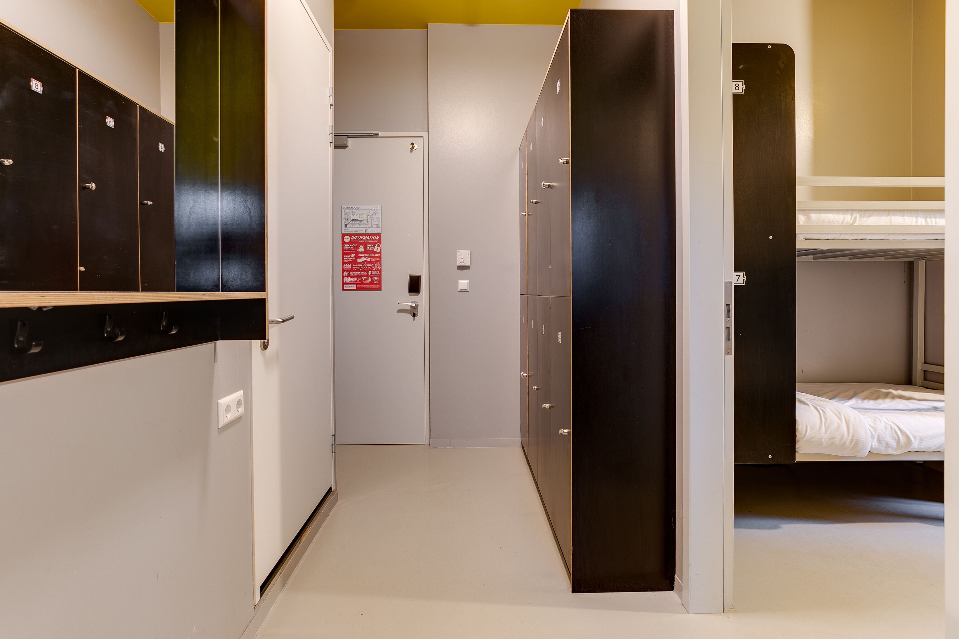 A corridor with lockers, a door with a sign, and a bunk bed area.