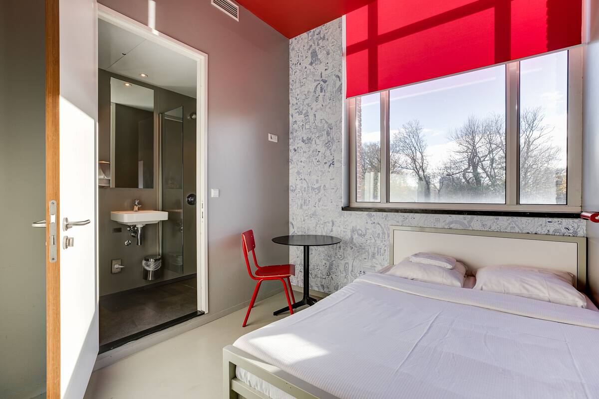 A modern bedroom with a bed, a red chair, a small table, and a window with a view of trees.
