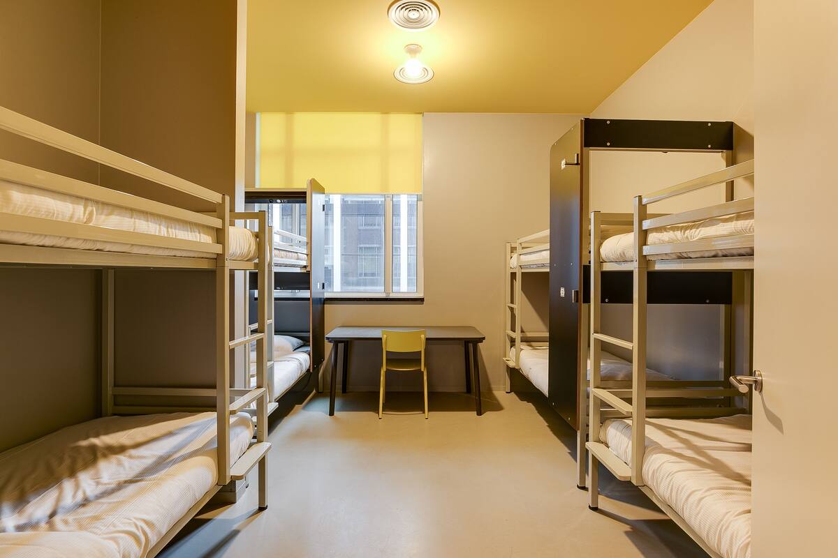 A dormitory room with bunk beds, a desk, and a window.