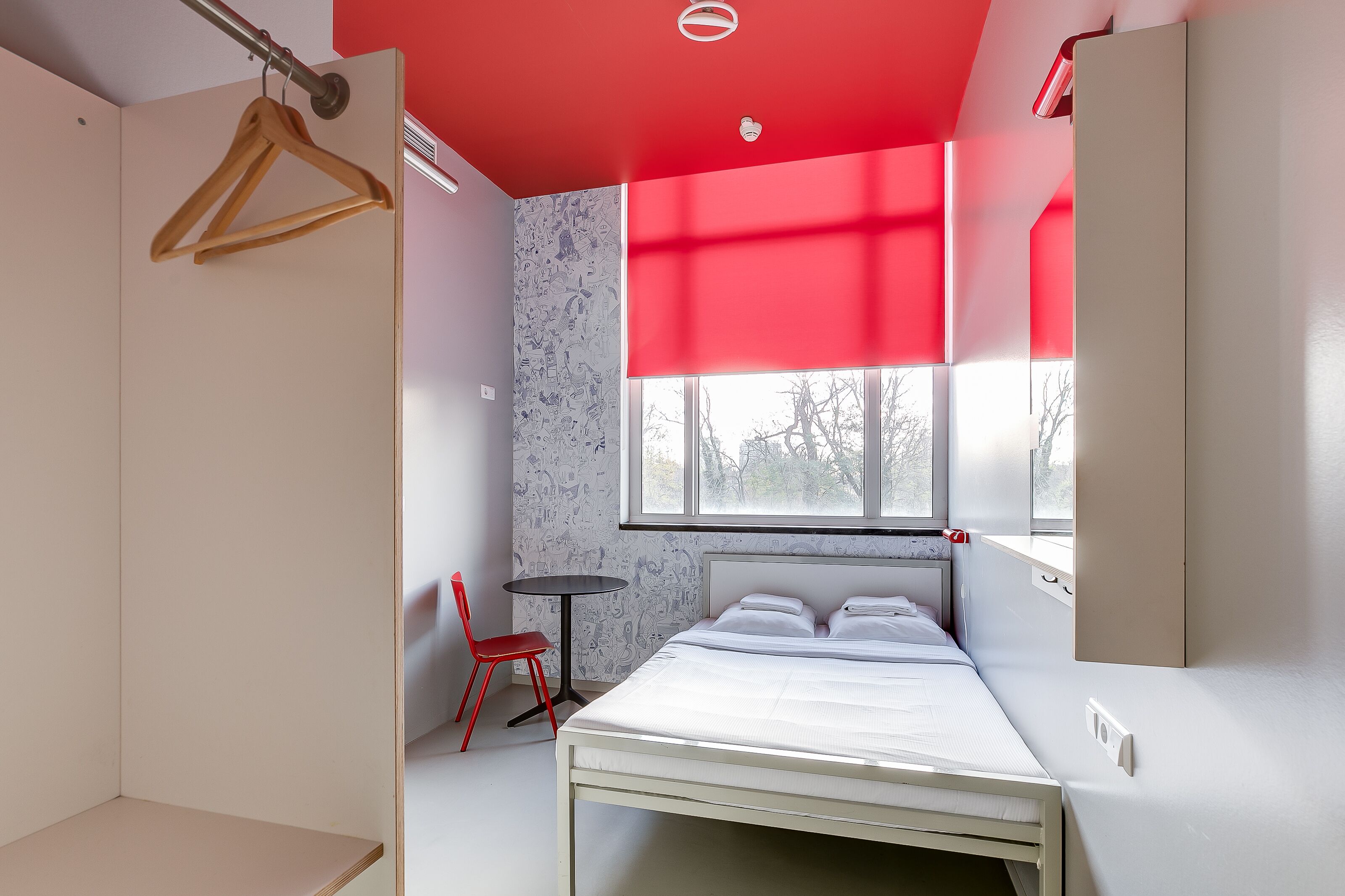 A modern bedroom with a bed, a red chair, a small table, and a window with blinds.