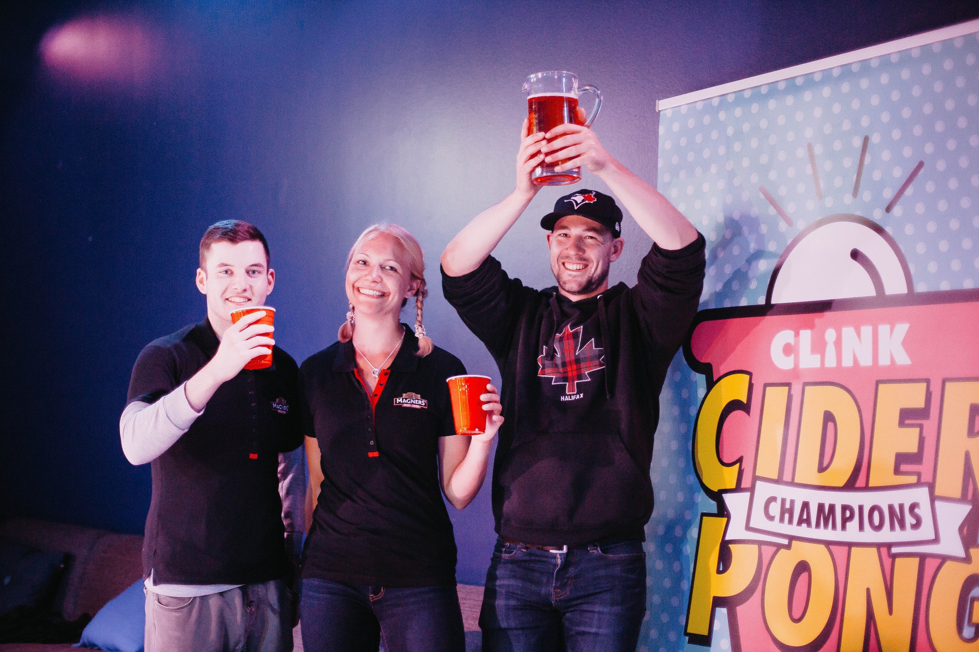 Drie personen met drankjes voor een bord met de tekst 'Clink Cider Champions'.