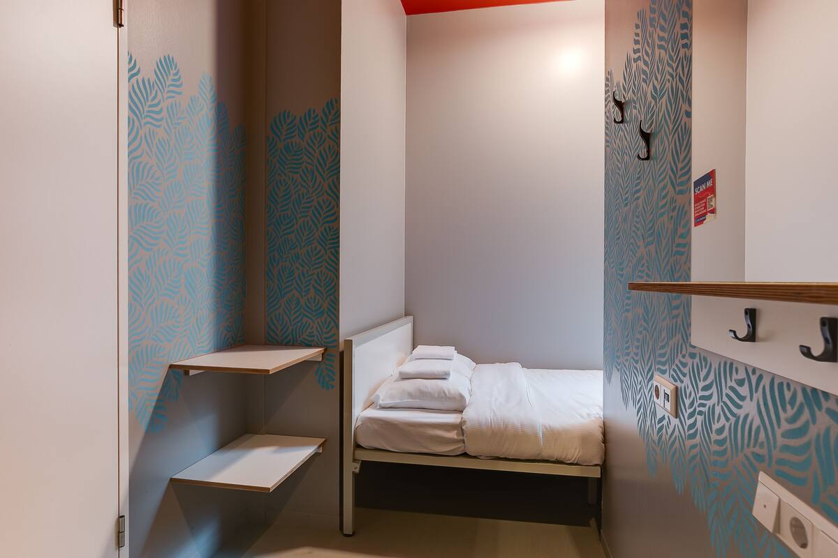 A small, compact room with a bed, a shelf, and a wall-mounted light switch.