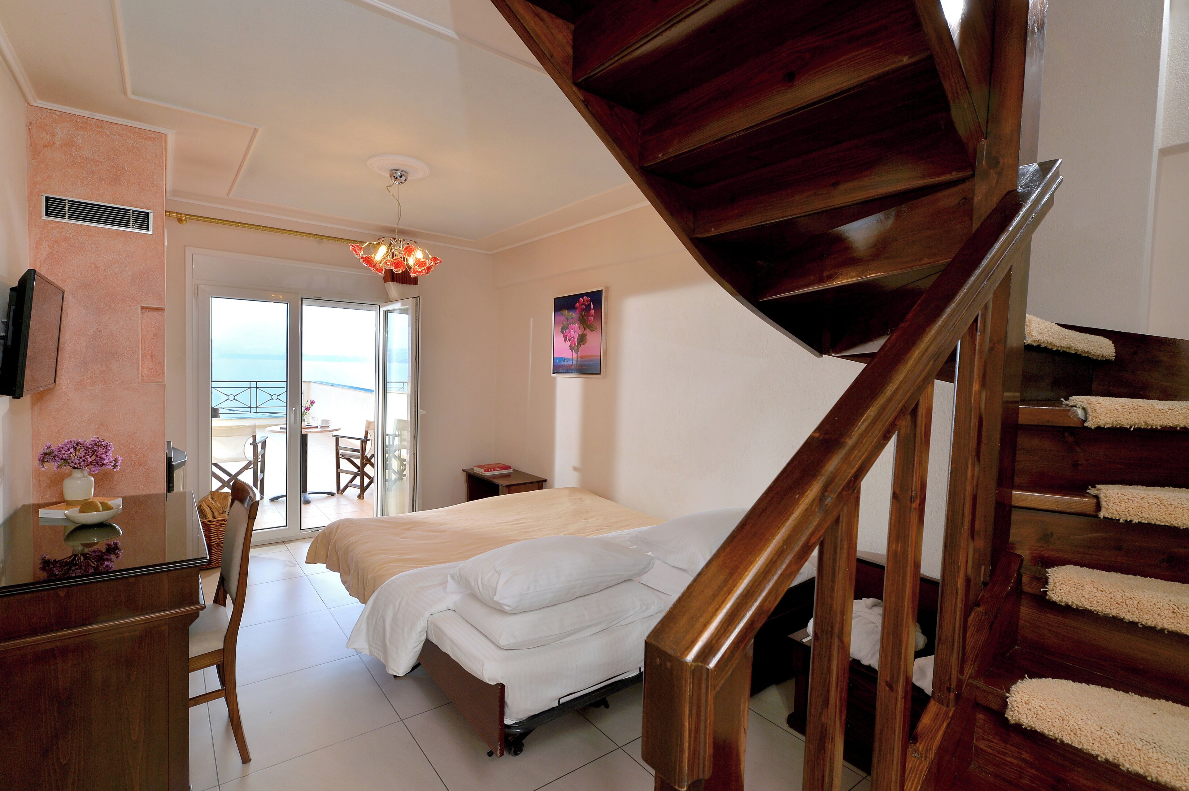 A bedroom with a bed, a desk, a chair, a wooden staircase, and a view of the outdoors.