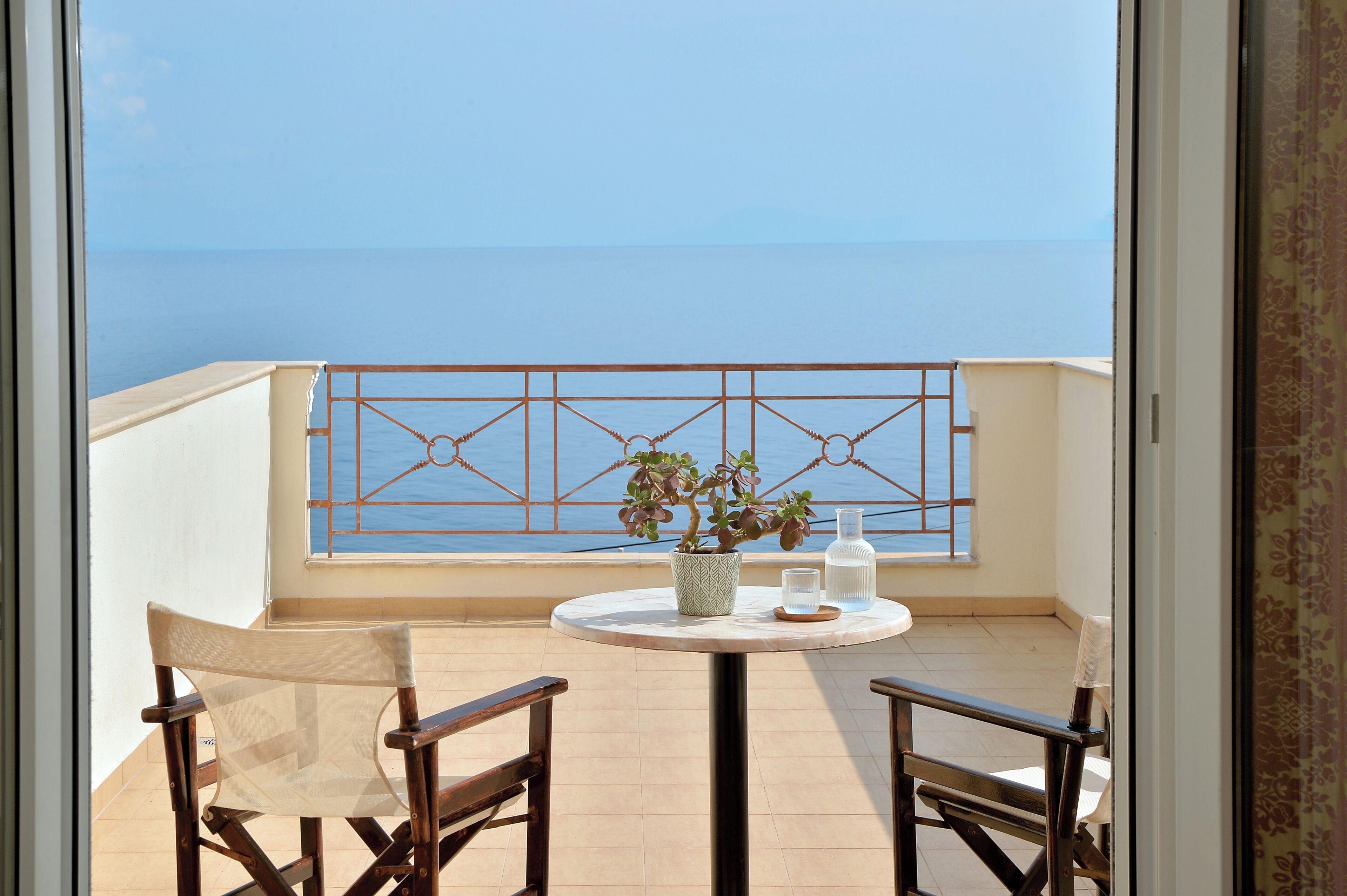 A balcony with a view of the sea, a round table with a plant, two chairs, and a metal railing.