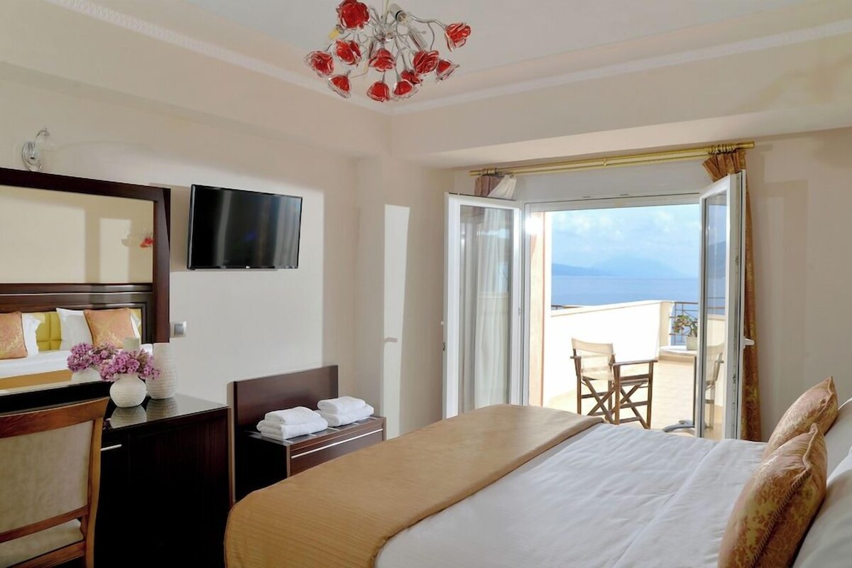 A hotel room with a bed, a TV, a desk, a chair, a balcony with a view, and a chandelier.