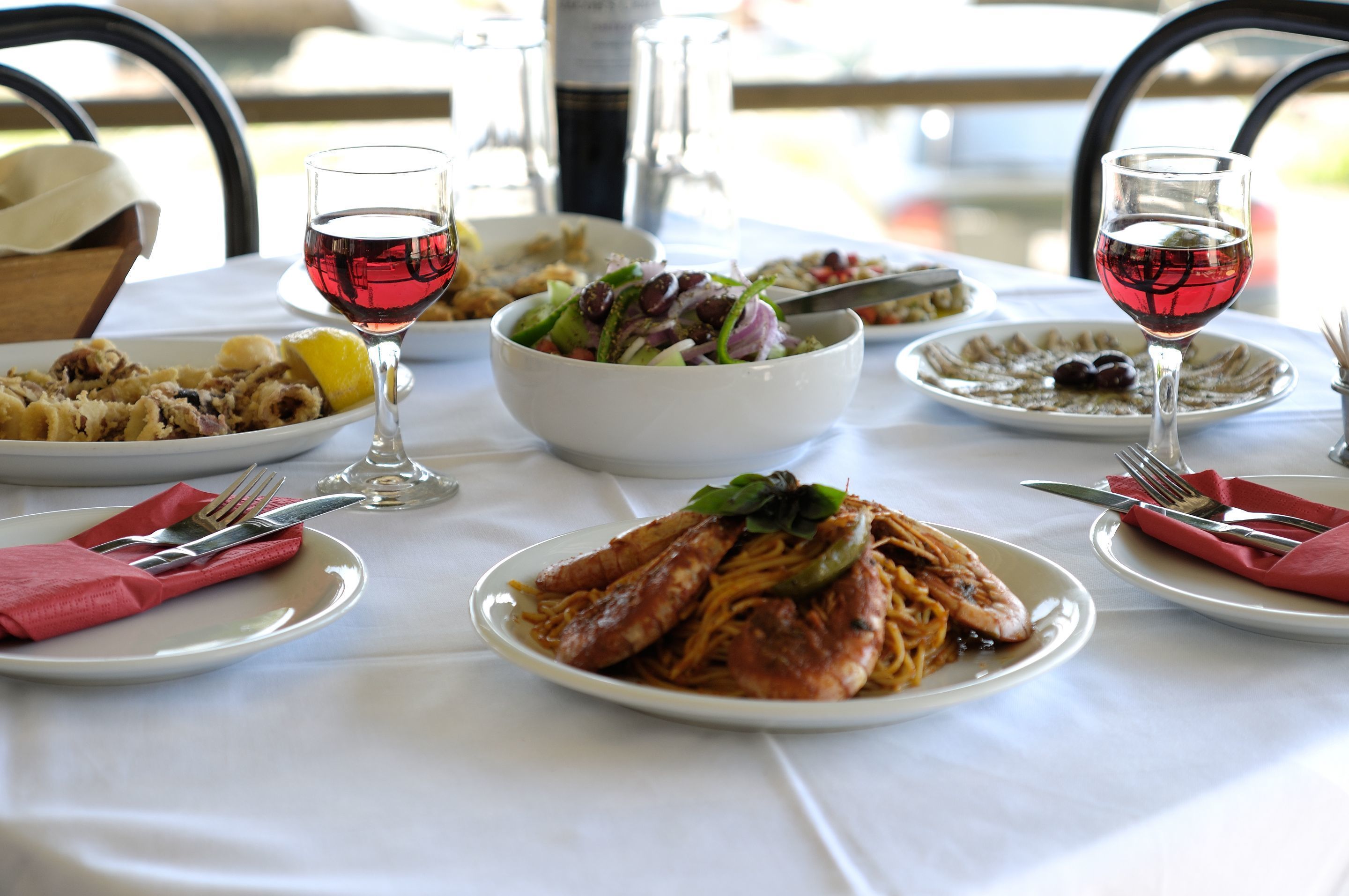 A table set with various dishes, including pasta and seafood, and two glasses of red wine.