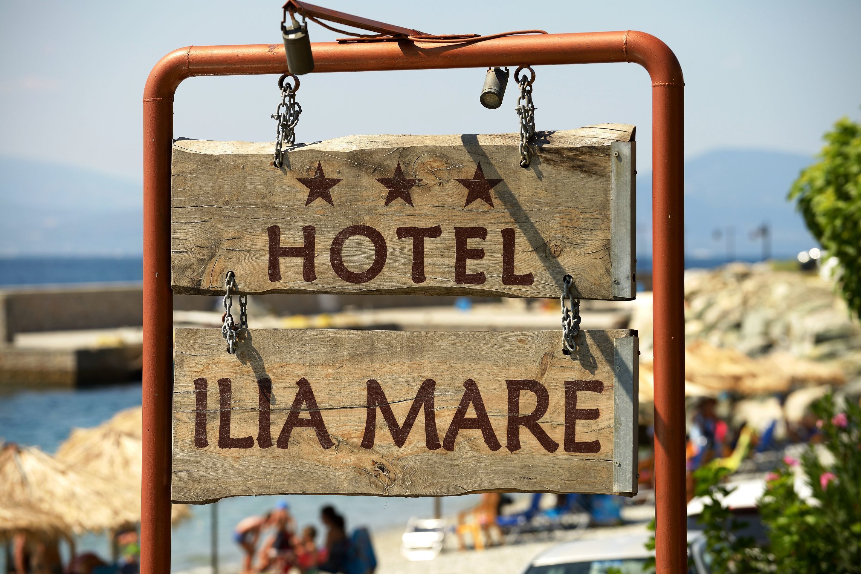 A wooden sign with "HOTEL" and "ILIA MARE" written on it, hanging from a metal frame.