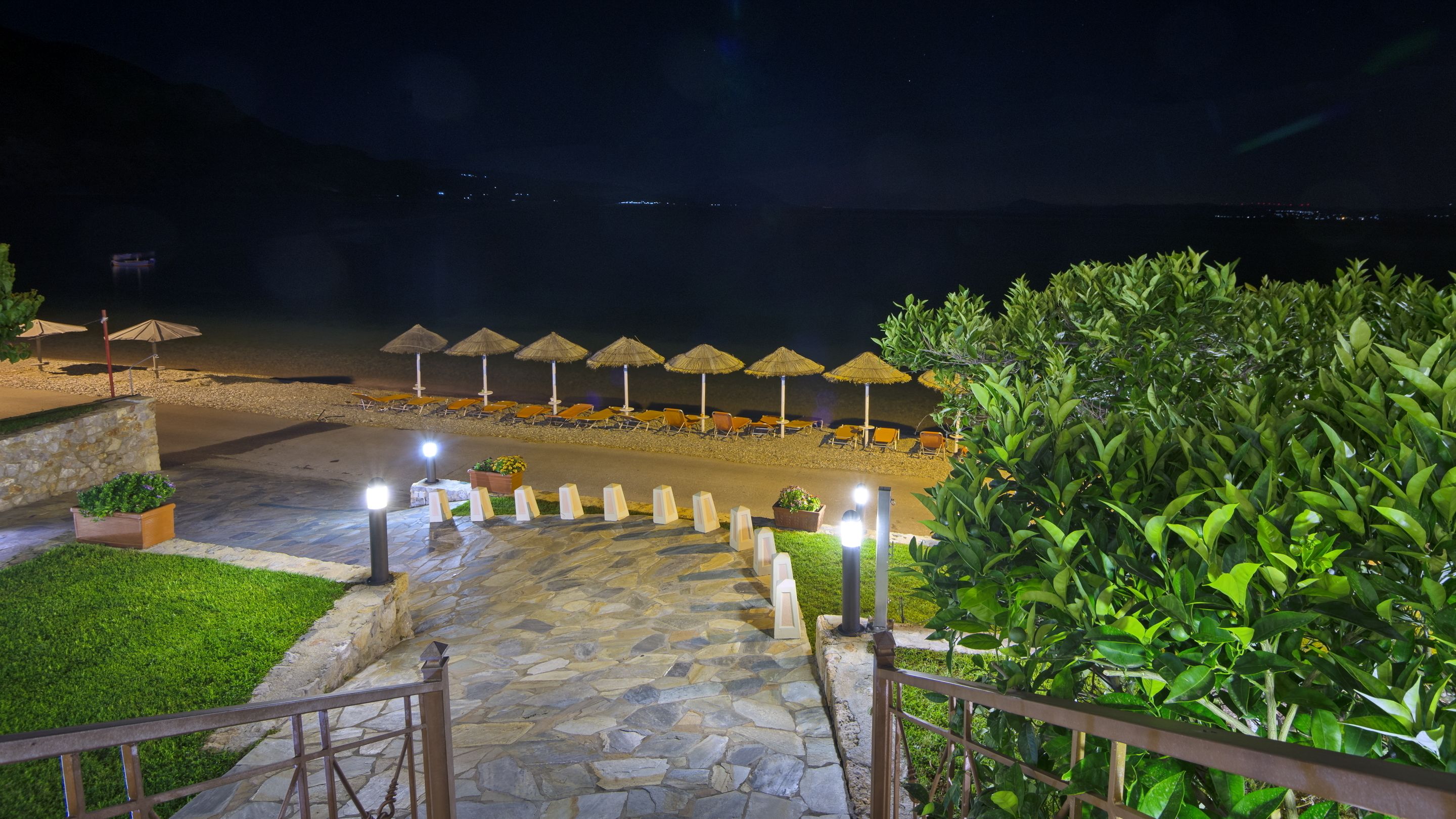 A stone pathway leading to beachside seating with umbrellas, a view of the sea, and a well-lit entrance.