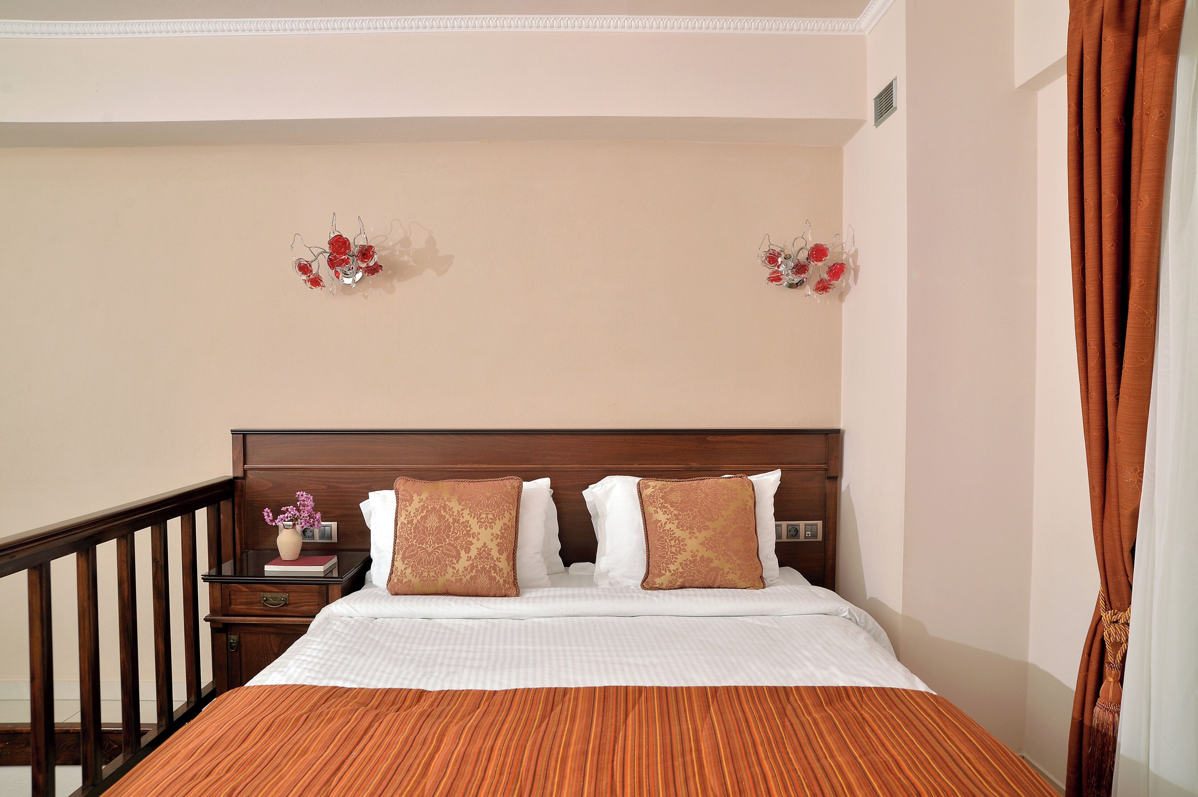 Habitación de hotel con cabecera de madera, cama con ropa blanca, dos apliques decorativos, mesita de noche con un jarrón de flores y cortina marrón.