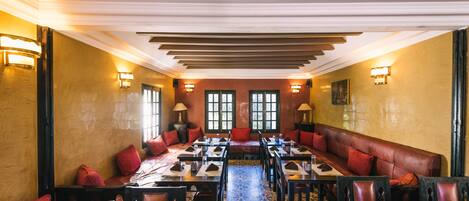 A dining area with a long table, multiple chairs, and a sofa. There are wall sconces and a large window with multiple panes.