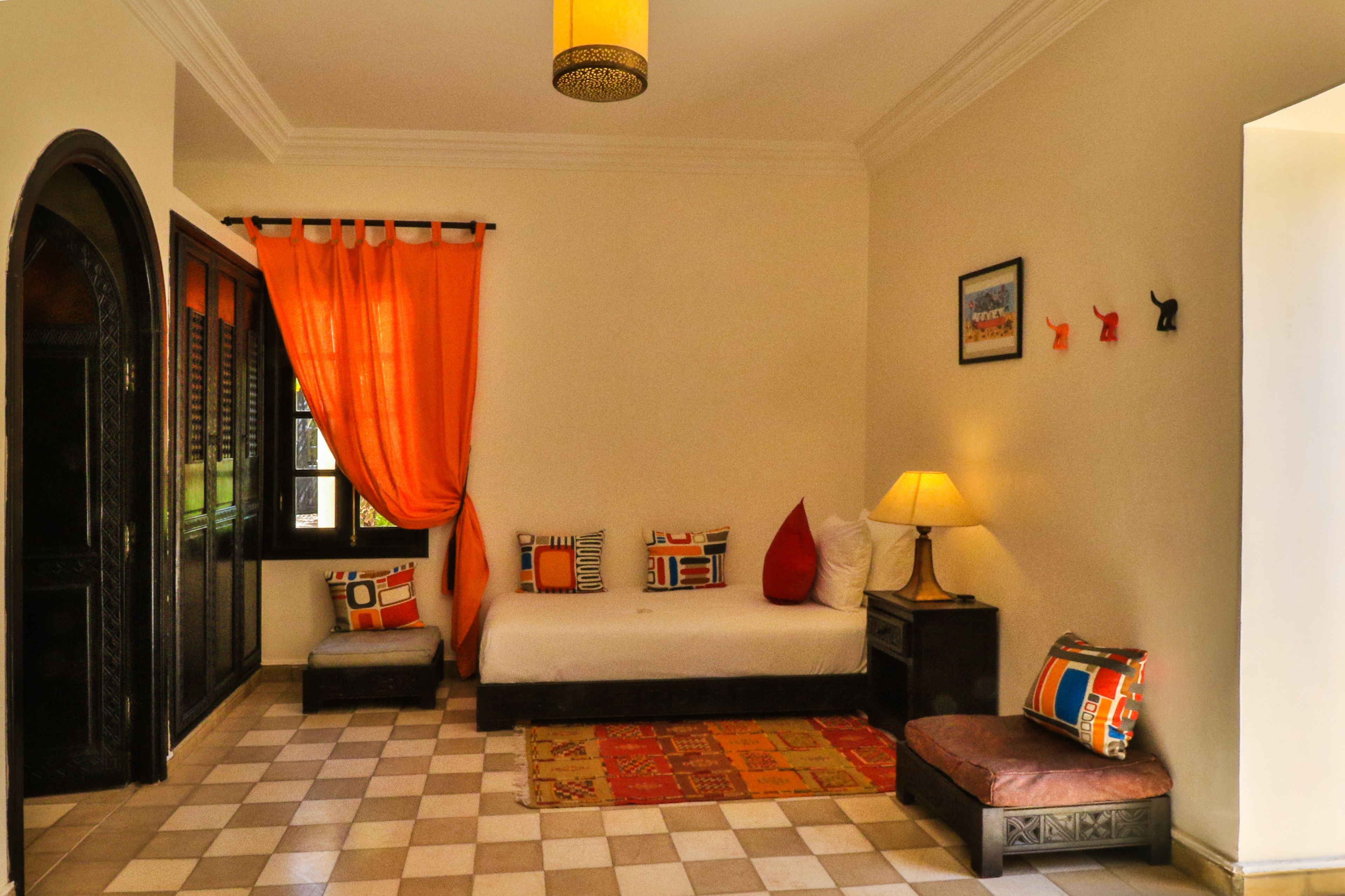 Quarto com piso xadrez, cama com várias almofadas, mesa de cabeceira com abajur e janela com cortinas laranja.